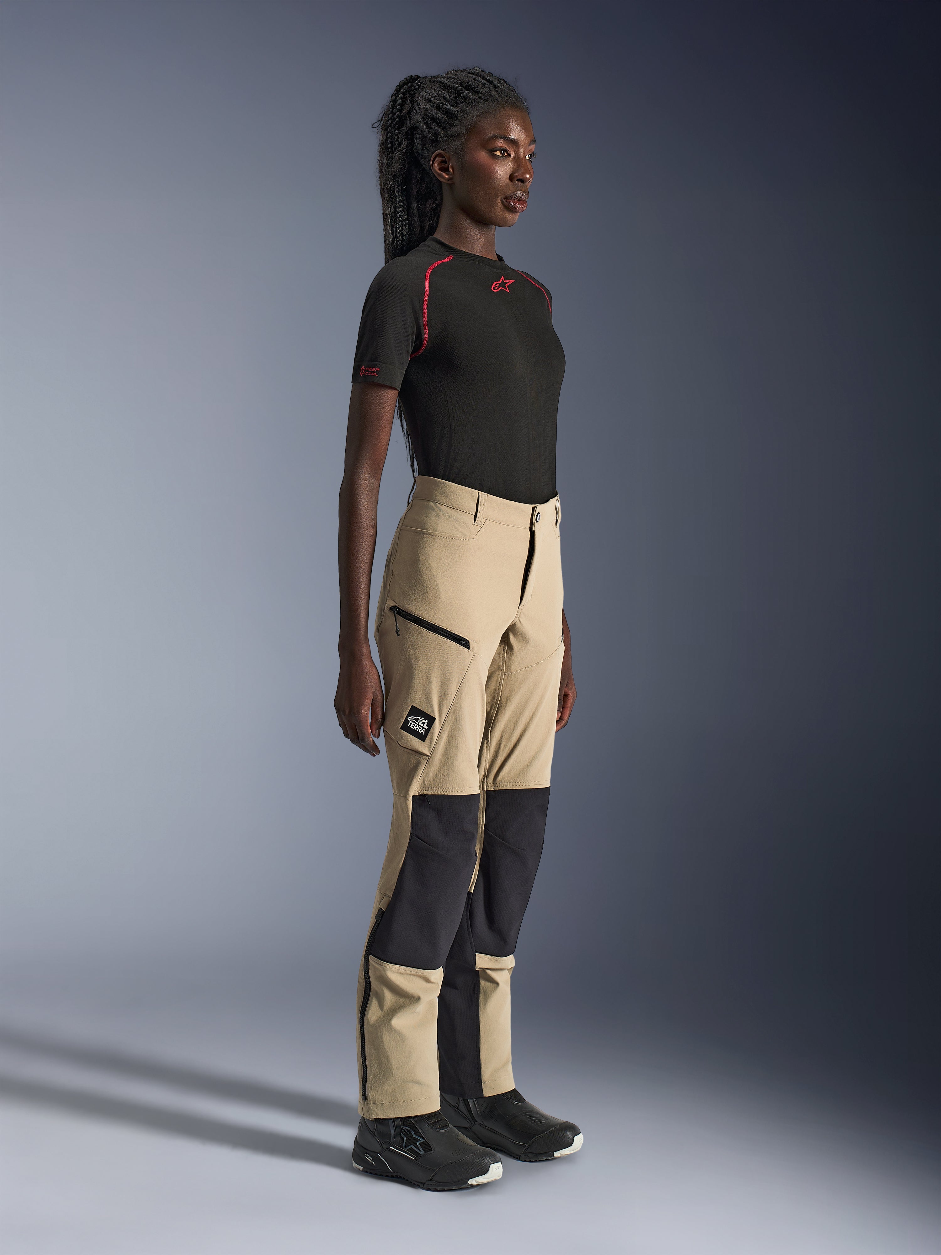 Calças Alpinestars Stella Flex-AST Explorer para Mulher, calças de aventura e touring em areia e preto, perfil lateral de uma modelo a usar uma t-shirt preta Alpinestars e botas de motociclismo, com reforços de ripstop nos joelhos e bolsos cargo com fecho de correr.