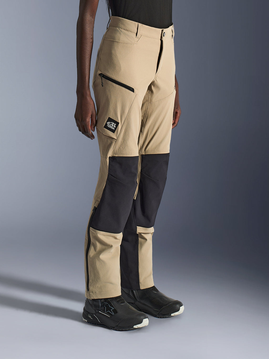 Mulher a usar calças Alpinestars Stella Flex-AST Explorer, calças de motociclismo de aventura em areia e preto com painéis de ripstop pretos nos joelhos, bolsos cargo com fecho de correr, emblema com o logótipo 'ALL TERRA' e fechos na parte inferior das pernas.