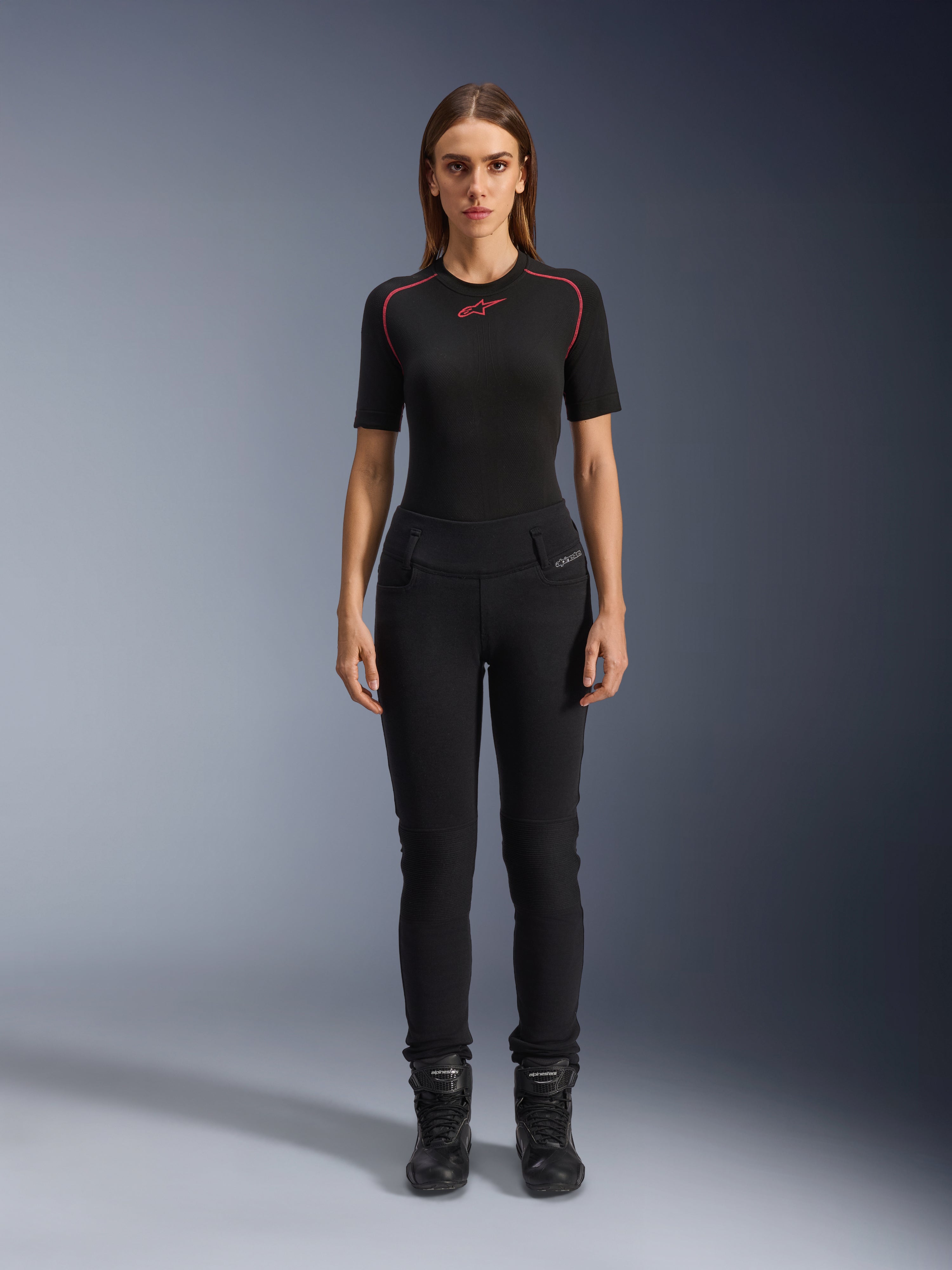 Alpinestars Womens Banshee Leggings, leggings de motociclismo pretas, ajuste anatómico que se molda ao corpo, usadas por uma modelo feminina com uma camada de base preta e botas de motociclismo, com painéis canelados nos joelhos e cintura subida para condução urbana.