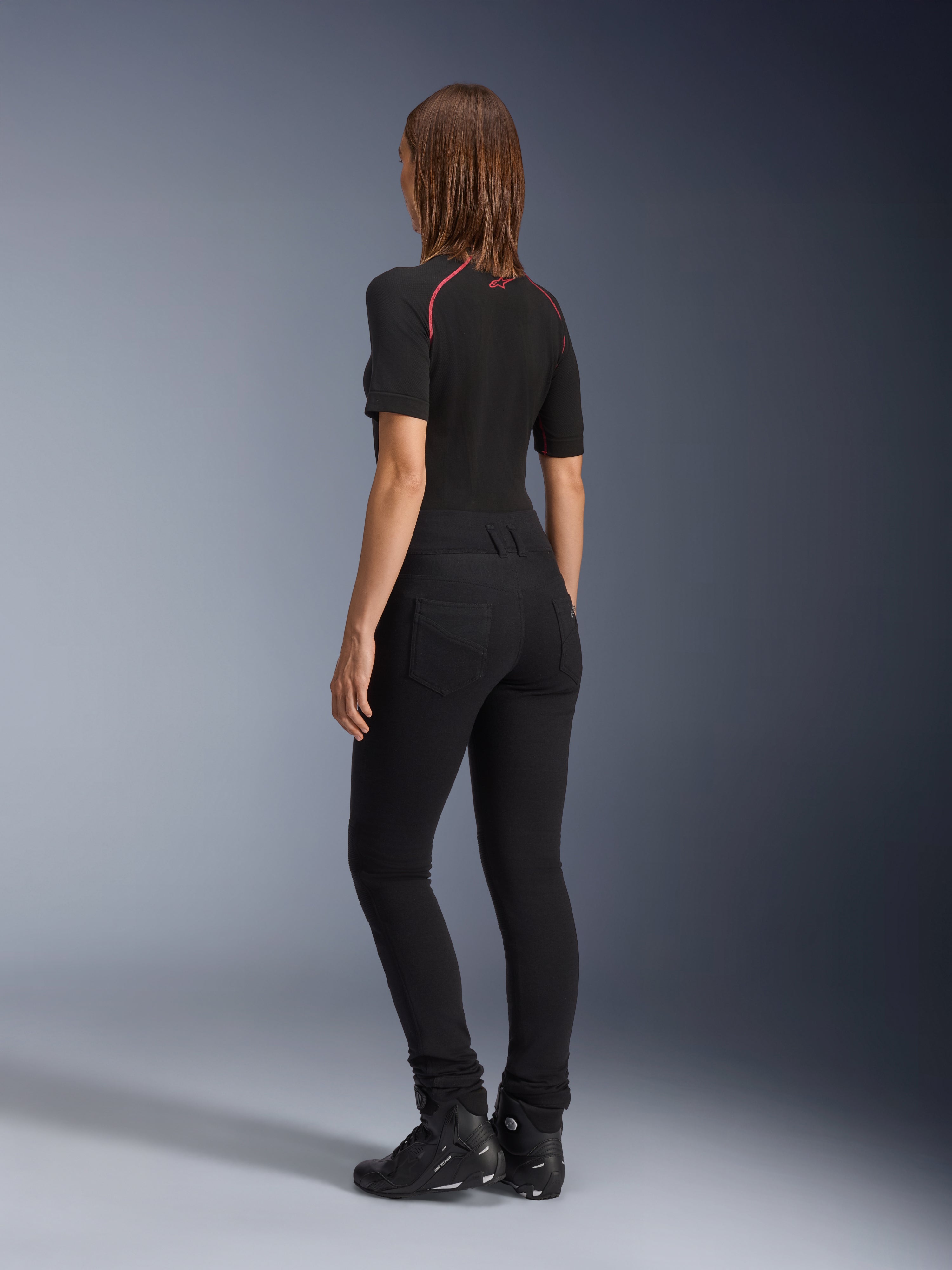 Alpinestars Leggings Banshee Femininas, pretas, vista traseira que mostra o ajuste anatómico elástico e os bolsos traseiros, usadas com um top preto de manga curta e botas de motociclismo, equipamento de motociclismo urbano elegante para mulher