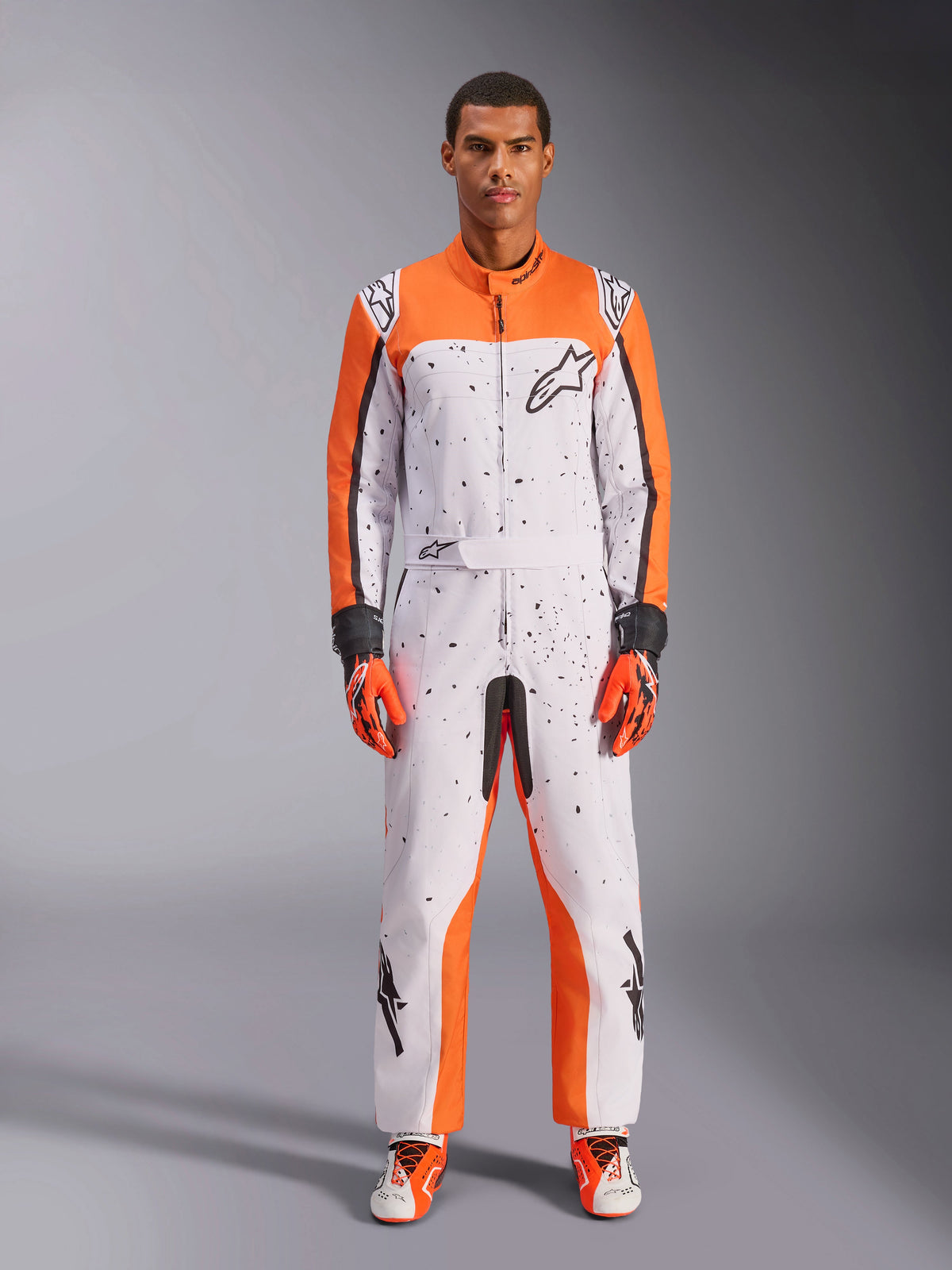 Macacão Alpinestars KMX-9 V3 Graphic 6, fato de karting, branco, laranja e preto, com um padrão salpicado a preto, usado com luvas e calçado de competição a condizer em laranja e preto, equipamento de proteção integral para desporto motorizado com logótipos destacados e design atlético