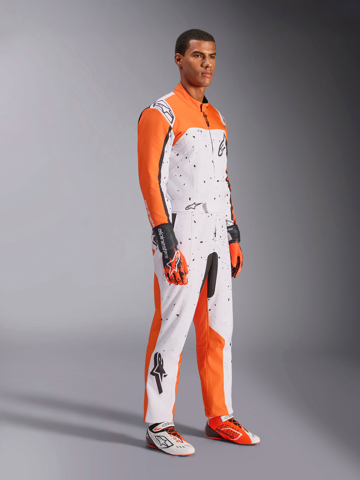 Macacão de Karting Alpinestars KMX-9 V3 Graphic 6, branco, laranja e preto, com um padrão salpicado e mangas laranja vibrantes, vestido por um modelo com luvas e botas de corrida a combinar, equipamento profissional de desporto motorizado de dupla camada.