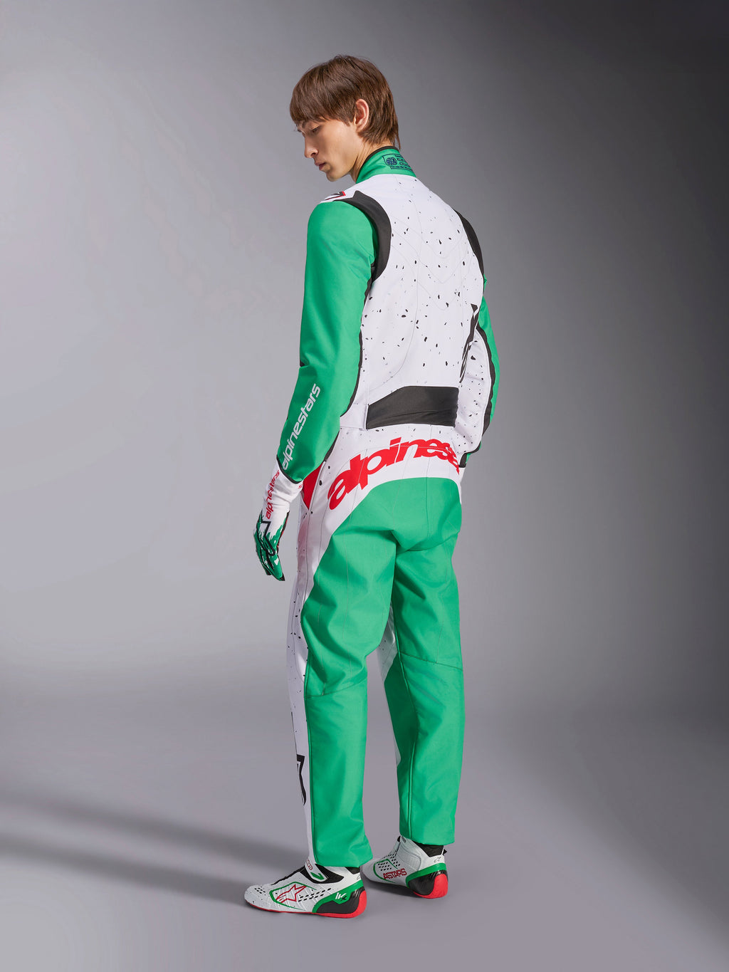 Fato Alpinestars KMX-9 V3 Graphic 6, fato de karting, branco, verde e vermelho, modelo visto de costas lateralmente, com mangas e painéis inferiores verdes, tronco superior branco com detalhes de salpicos pretos, logótipo Alpinestars em vermelho na zona lombar, acompanhado por sapatos de karting brancos e verdes a condizer