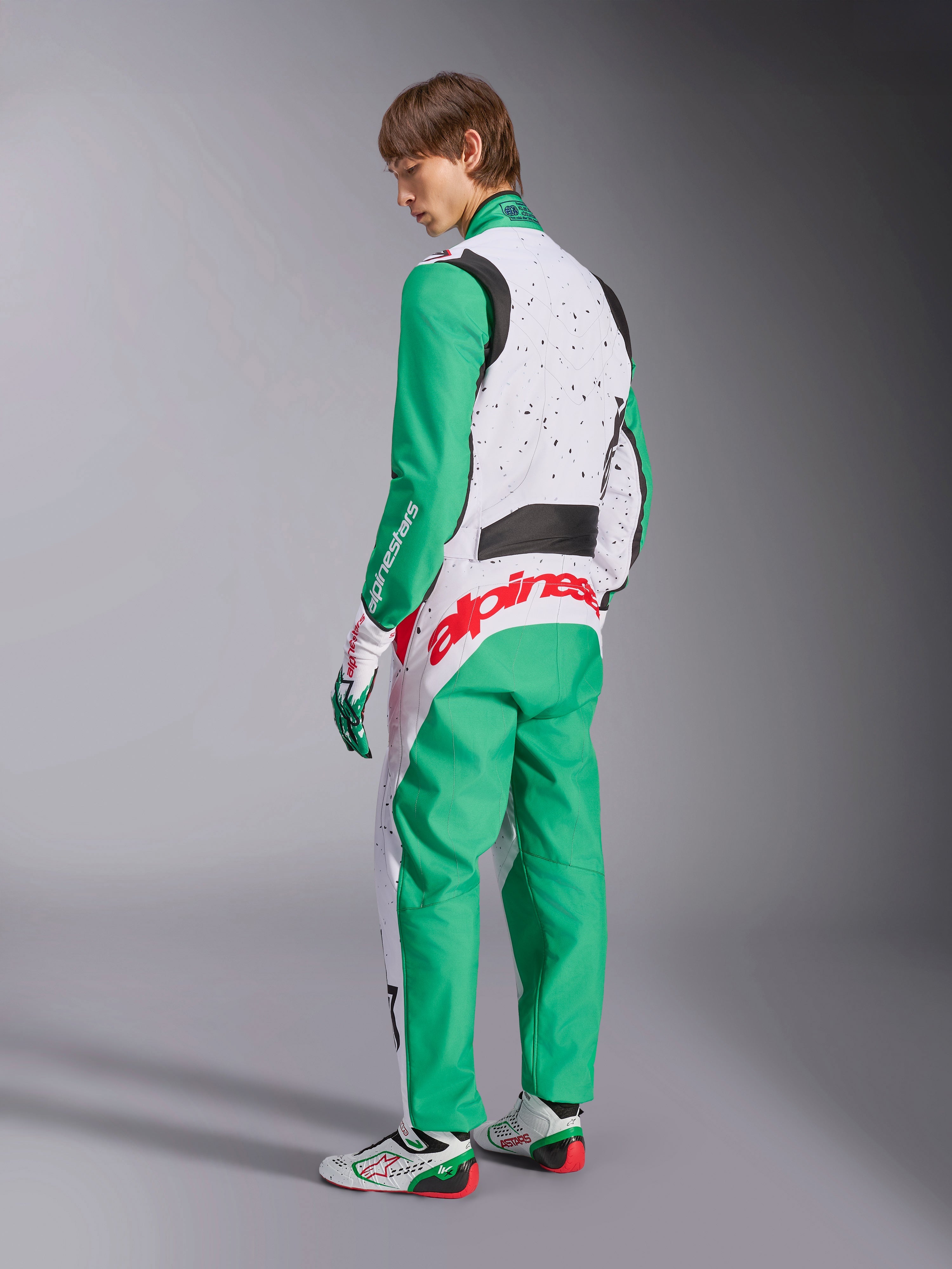 Fato Alpinestars KMX-9 V3 Graphic 6, fato de karting, branco, verde e vermelho, modelo visto de costas lateralmente, com mangas e painéis inferiores verdes, tronco superior branco com detalhes de salpicos pretos, logótipo Alpinestars em vermelho na zona lombar, acompanhado por sapatos de karting brancos e verdes a condizer