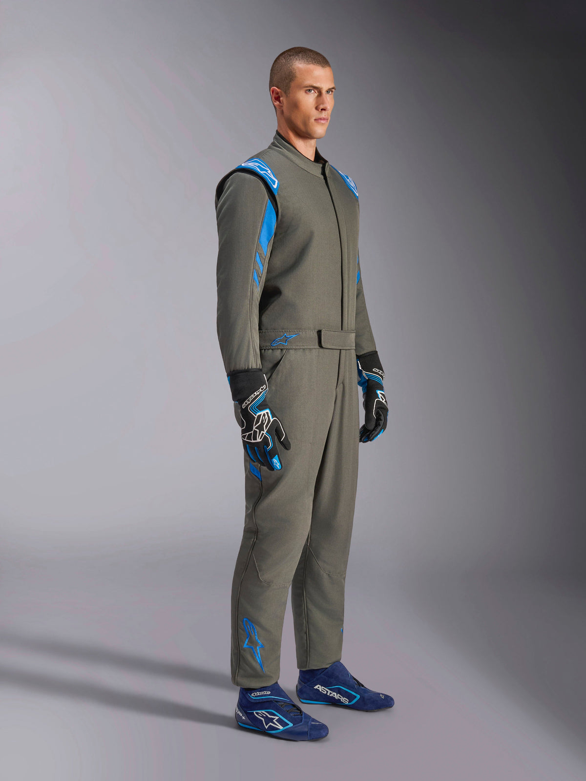 Fato Alpinestars GP Race V3 FIA em Anthracite Blue, fato de competição automóvel de peça única usado por um modelo masculino, apresenta detalhes em azul brilhante nos ombros e tornozelos, combinado com luvas de corrida coordenadas e sapatos de corrida azuis, equipamento profissional de desporto motorizado