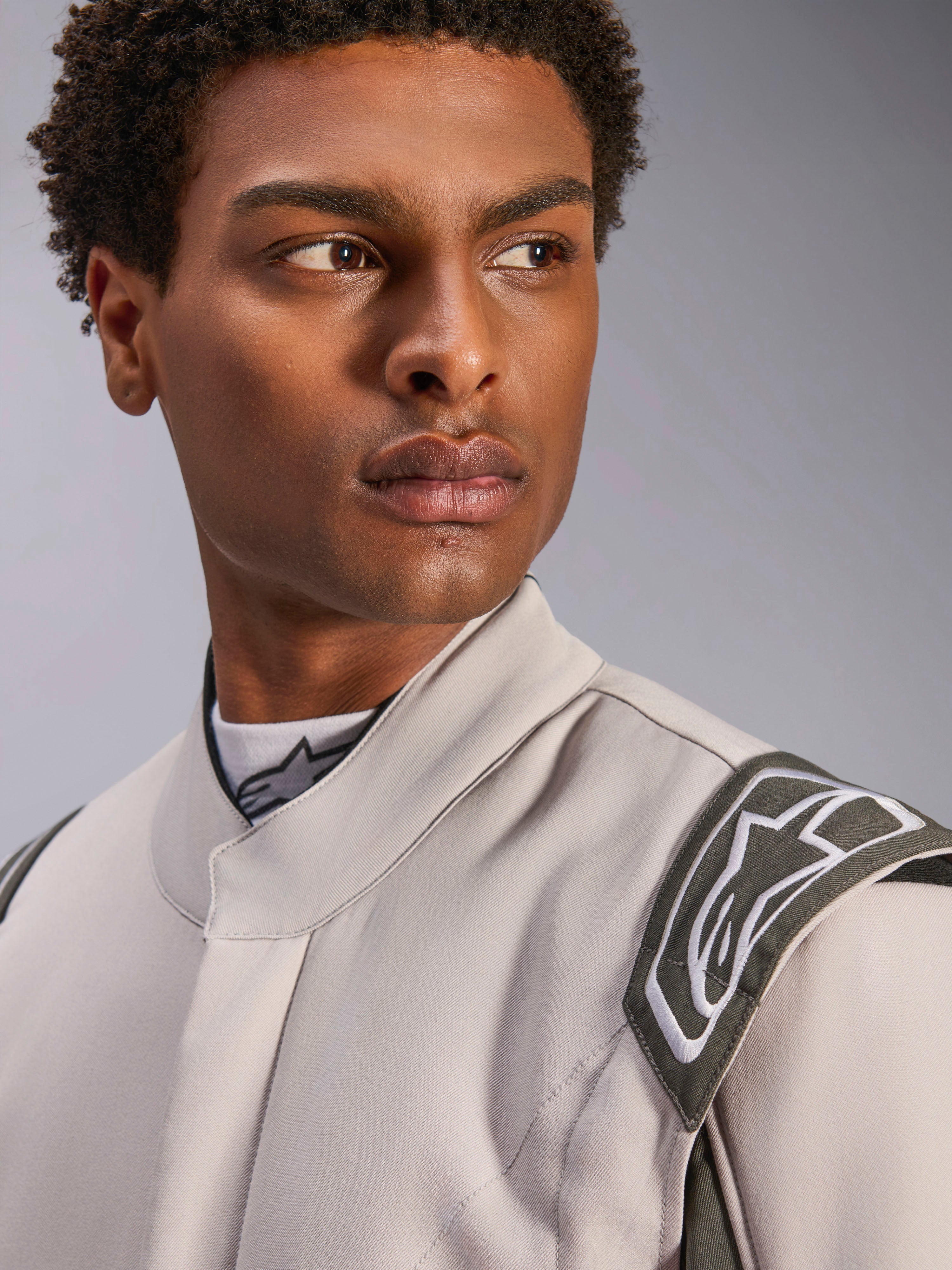 Modelo a usar o fato Alpinestars GP Race V3 FIA em Silver Anthracite, plano aproximado focado no tronco superior e no ombro, destacando a epaulette de extração e o colarinho ergonómico, equipamento profissional de automobilismo