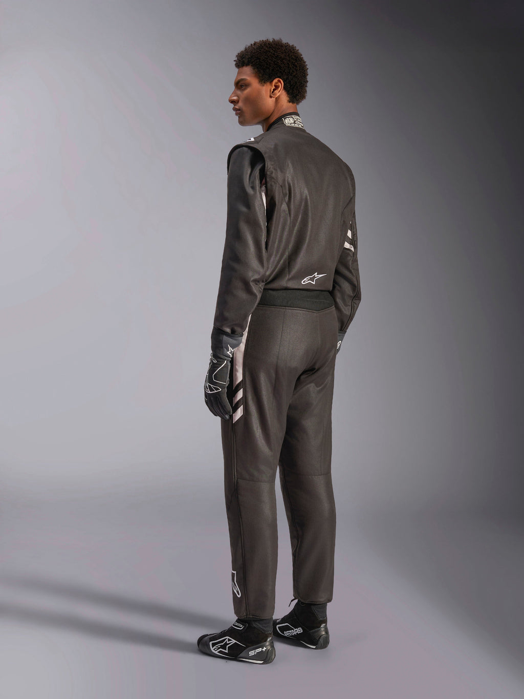 Alpinestars GP Race V3 Suit FIA, fato de automobilismo preto com detalhes prateados, usado por um modelo numa vista traseira de três quartos, combinado com luvas e botas de corrida pretas a condizer, equipamento profissional de desporto motorizado com um design elegante de duas camadas.