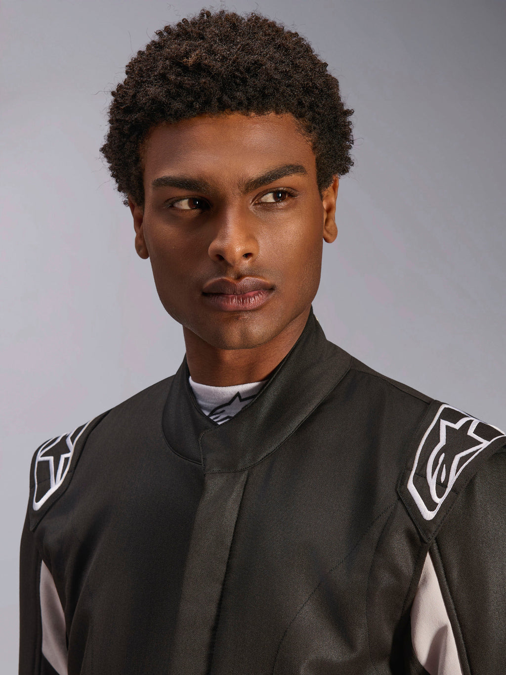 Macacão Alpinestars GP Race V3 FIA, fato de automobilismo preto usado por um homem, vista de perto com foco no colarinho macio e logótipos de estrela brancos nas ombreiras, equipamento de proteção profissional para desportos motorizados