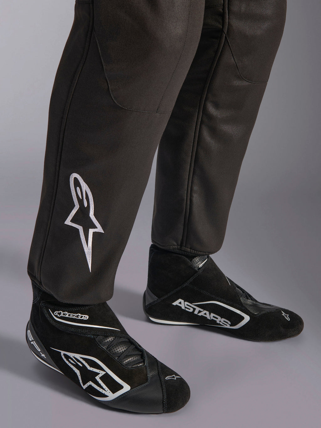 Macacão Alpinestars GP Race V3 FIA, grande plano da parte inferior das pernas e botas de competição, preto com detalhes prateados, com logótipo de estrela bordado a branco na canela e punhos canelados nos tornozelos, equipamento de proteção profissional para automobilismo