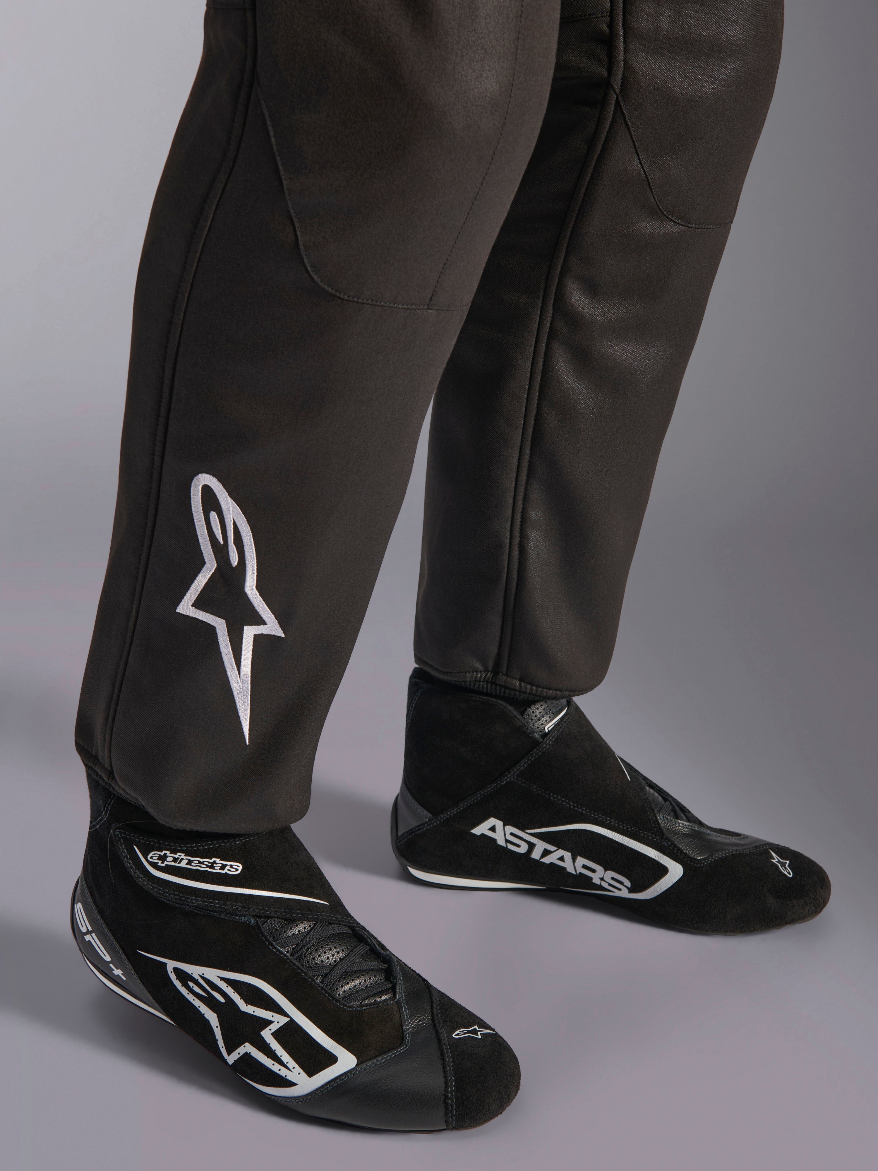 Macacão Alpinestars GP Race V3 FIA, grande plano da parte inferior das pernas e botas de competição, preto com detalhes prateados, com logótipo de estrela bordado a branco na canela e punhos canelados nos tornozelos, equipamento de proteção profissional para automobilismo