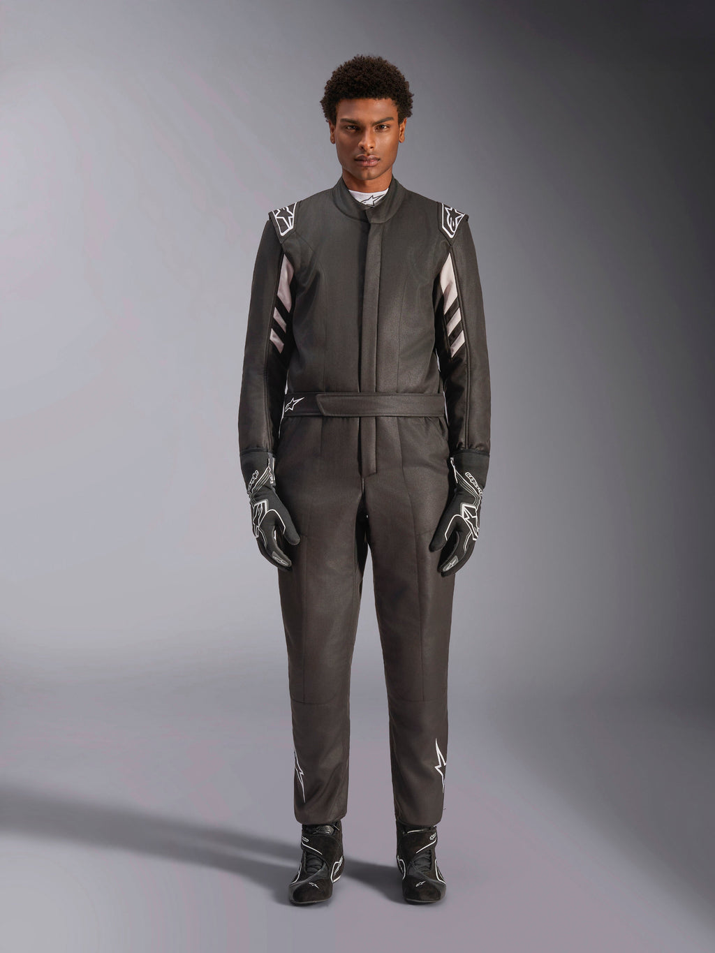Macacão Alpinestars GP Race V3 FIA, fato de automobilismo, preto e prateado, vista frontal de um piloto a usar equipamento de corrida completo, incluindo luvas e botas, design profissional de desporto motorizado com a marca Alpinestars nos ombros e no cinto