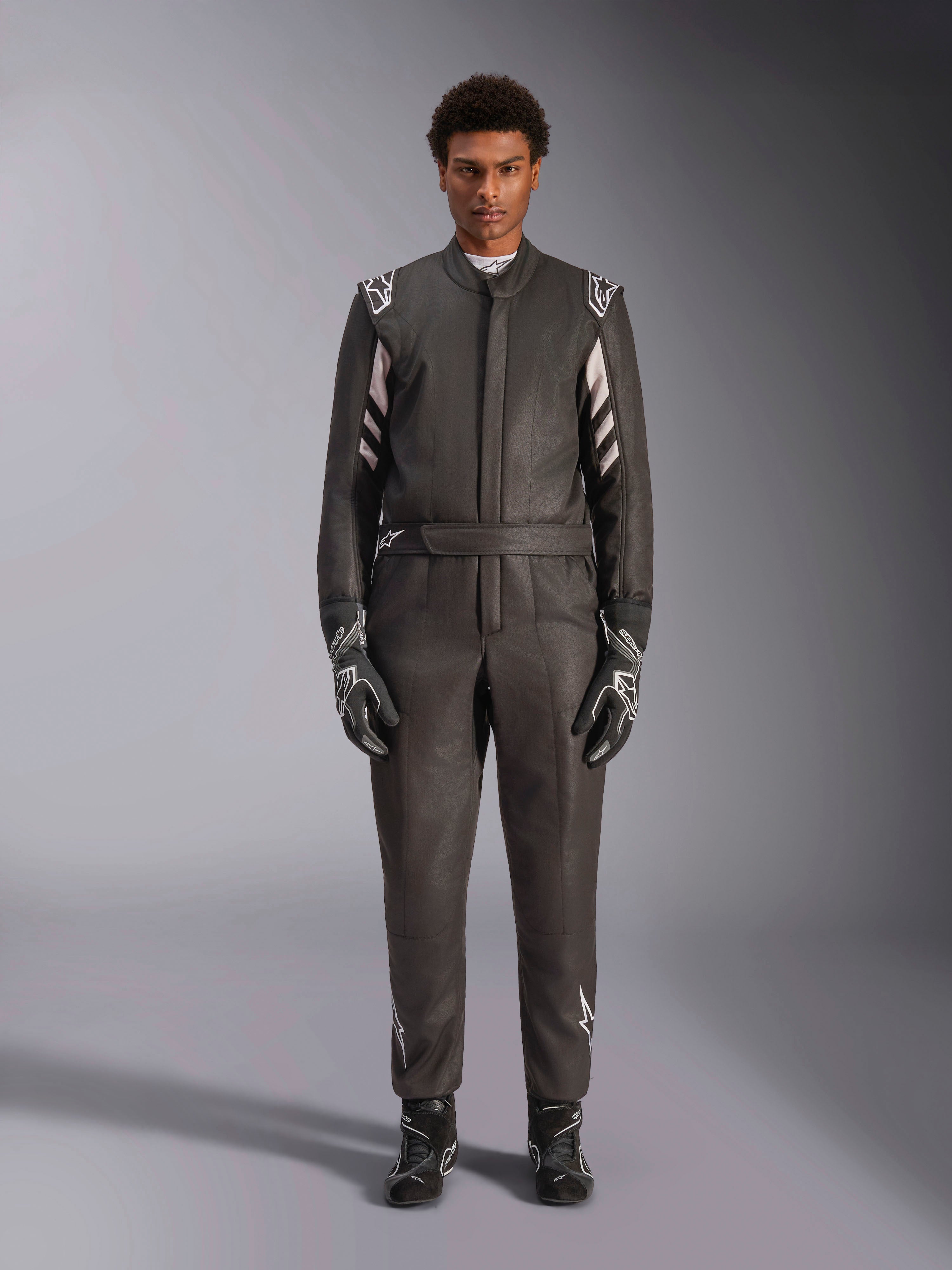 Macacão Alpinestars GP Race V3 FIA, fato de automobilismo, preto e prateado, vista frontal de um piloto a usar equipamento de corrida completo, incluindo luvas e botas, design profissional de desporto motorizado com a marca Alpinestars nos ombros e no cinto