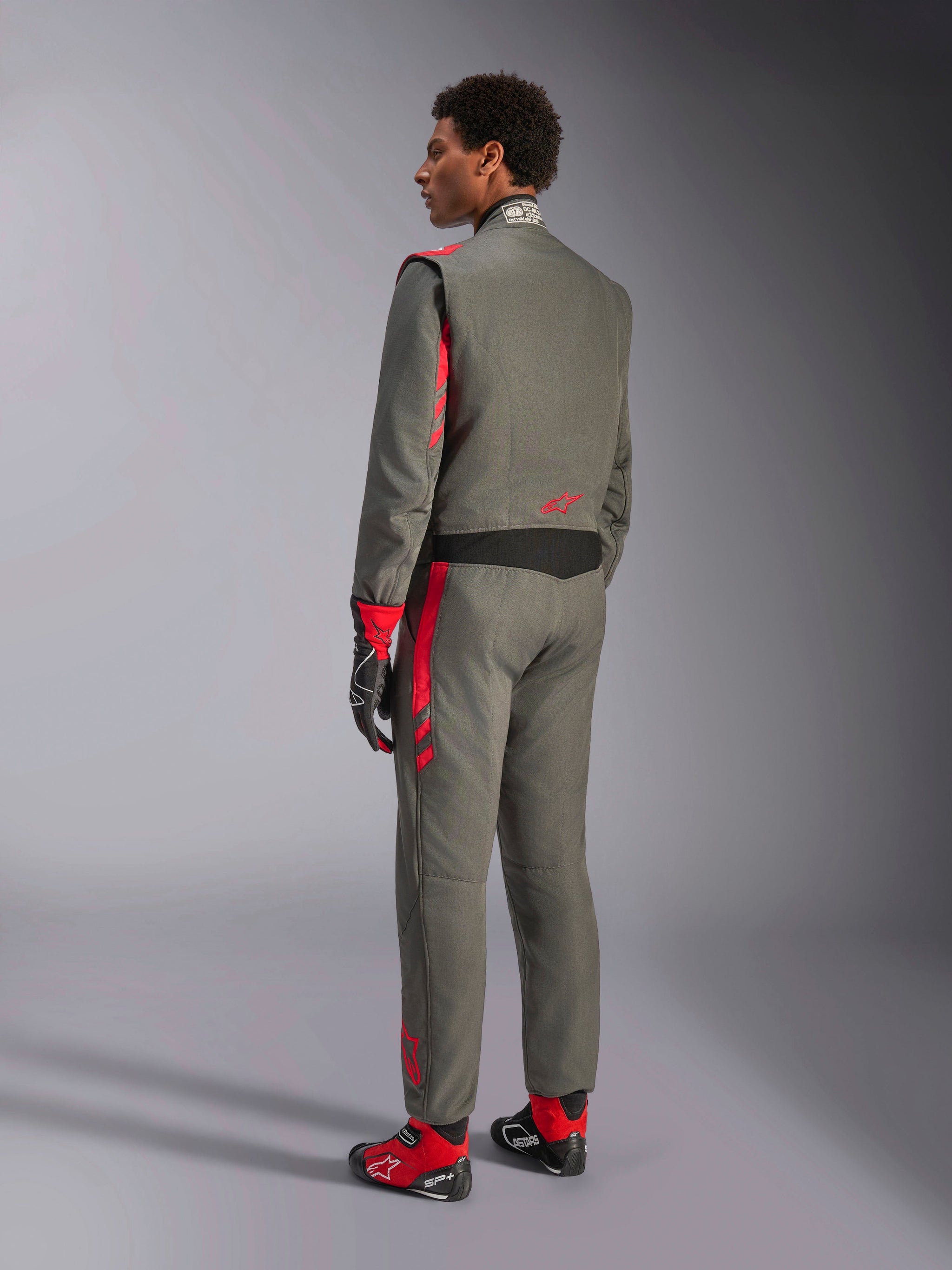 Alpinestars GP Race V3 Suit FIA, fato de corrida em Antracite Vermelho, vista traseira de modelo a usar o fato de duas camadas, detalhes vermelhos nos ombros e pernas, combinado com luvas e botas a condizer, equipamento profissional de automobilismo