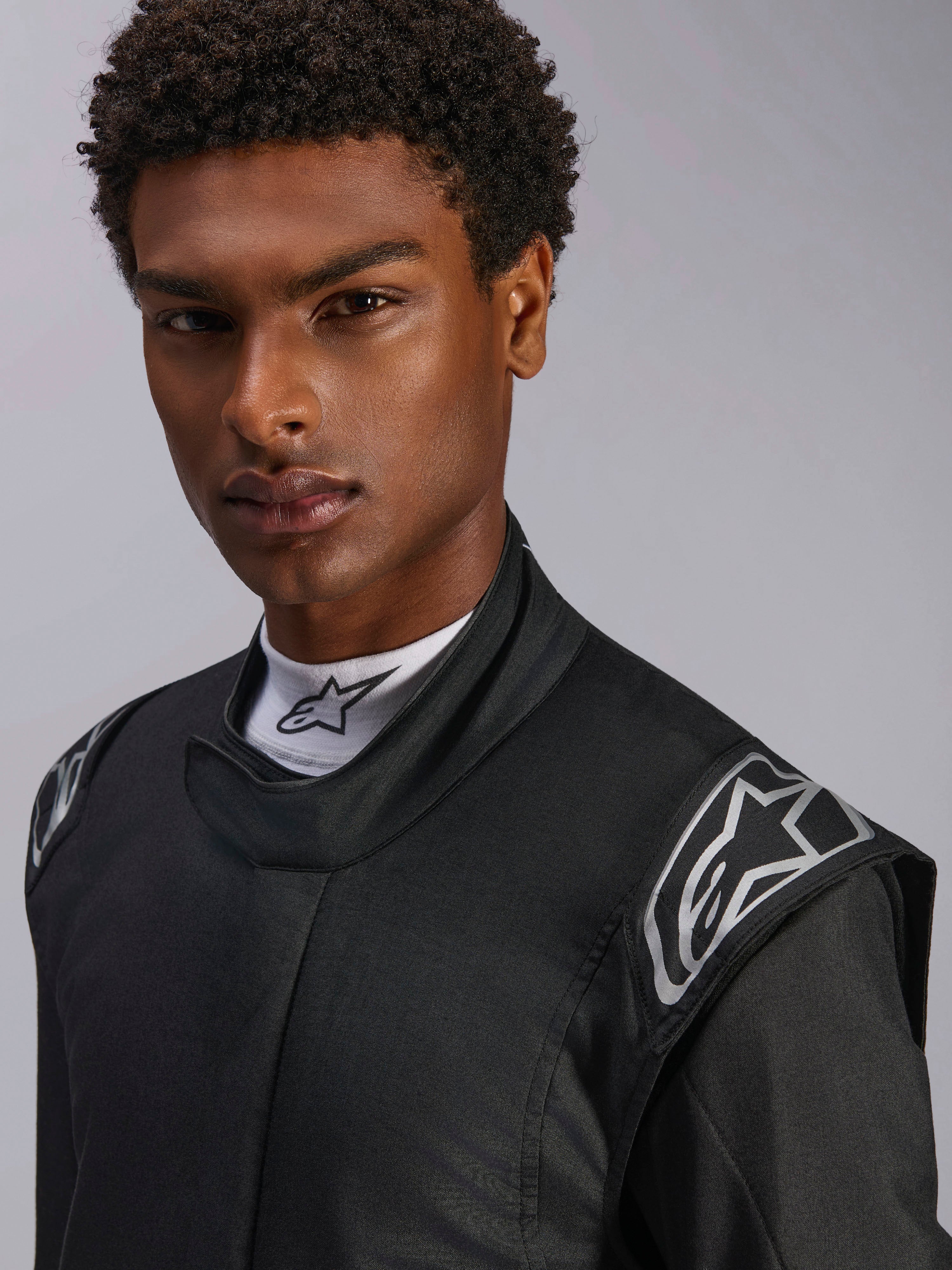 Alpinestars Hypertech V3 Suit, Black Dark Shadow auto racing suit, close-up on male model, silver shoulder logos, high collar with white Alpinestars mock neck shirt, professional motorsport protective gear
