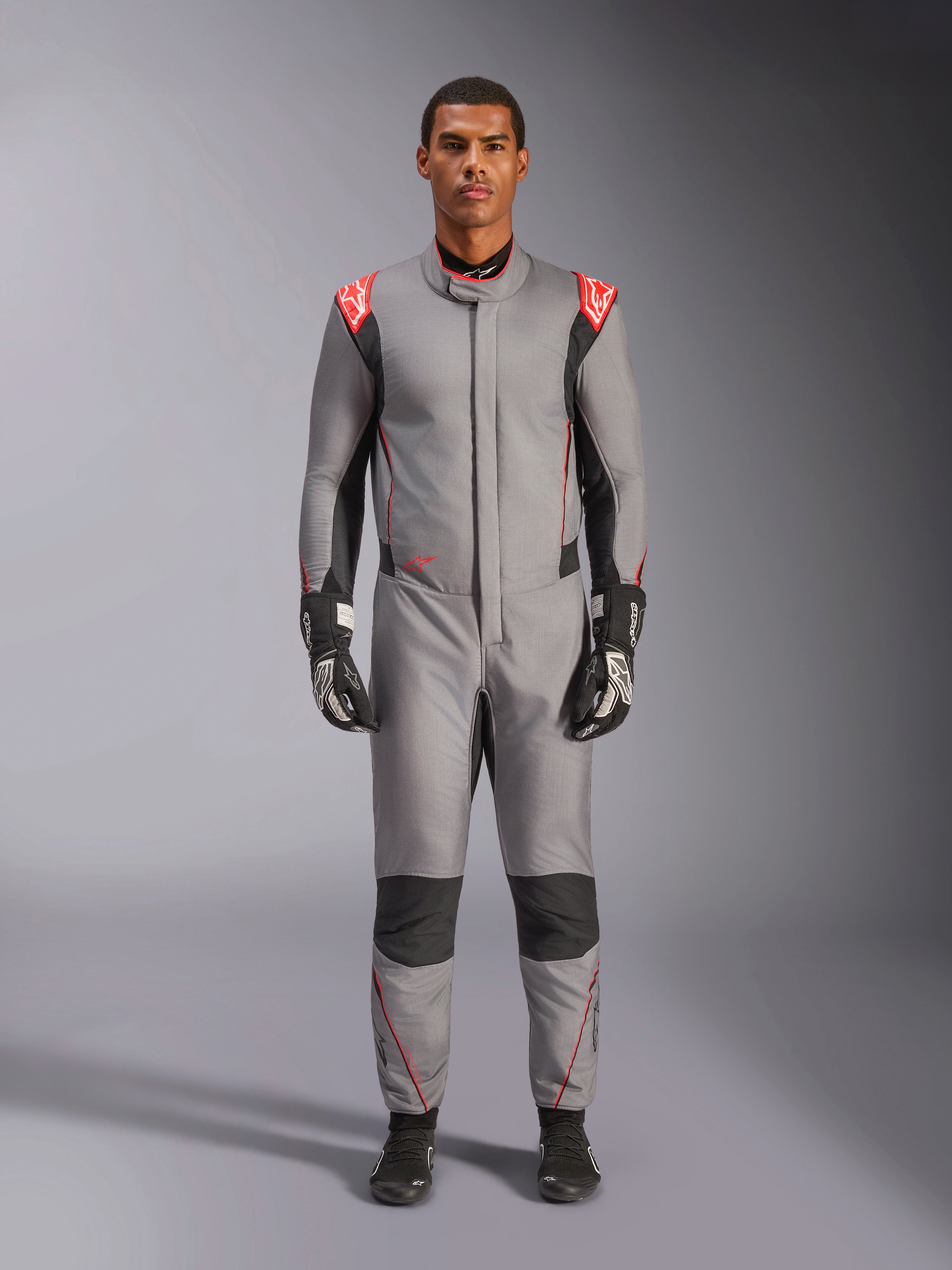 Alpinestars Hypertech V3 Suit, fato de automobilismo profissional em cinzento médio, preto e vermelho fluo, usado por um modelo com luvas e sapatos de corrida pretos, com um corte atlético ajustado, detalhes nos ombros em vermelho fluo e painéis pretos nos joelhos e laterais.