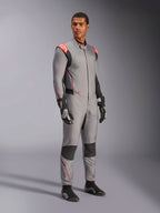 Male model wearing the Alpinestars Hypertech V3 Suit in Mid Gray with Black and Red Fluo accents, professional auto racing suit paired with black racing gloves and shoes, featuring a lightweight two-layer Nomex construction with red shoulder detailing and an athletic fit