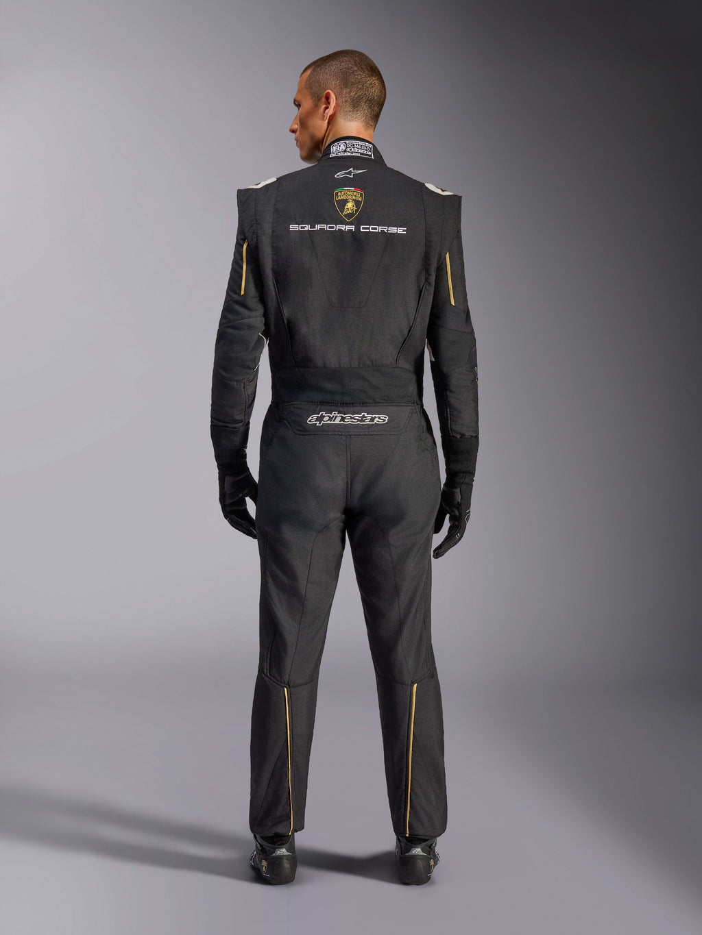 Alpinestars Limited Edition Lamborghini GP Tech V4 Suit FIA, Auto Racing Suit, Black with Gold and Silver accents, rear view showing Lamborghini Squadra Corse branding, Alpinestars logo, and gold piping on sleeves and legs, professional racing gear