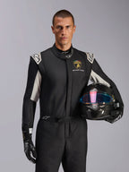 Male model wearing Limited Edition Lamborghini GP Tech V4 Suit, black racing suit with silver accents and Lamborghini Squadra Corse logo, holding a carbon fiber helmet, black racing gloves with white logos, professional auto racing gear
