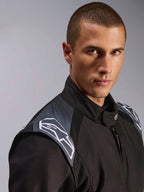 Modelo masculino a usar o Alpinestars KMX-5 V3 Suit, macacão de karting Black Dark Shadow, vista de perto da zona do peito e dos ombros, com detalhes cinzentos e logótipos Astars brancos, equipamento de proteção profissional para desportos motorizados