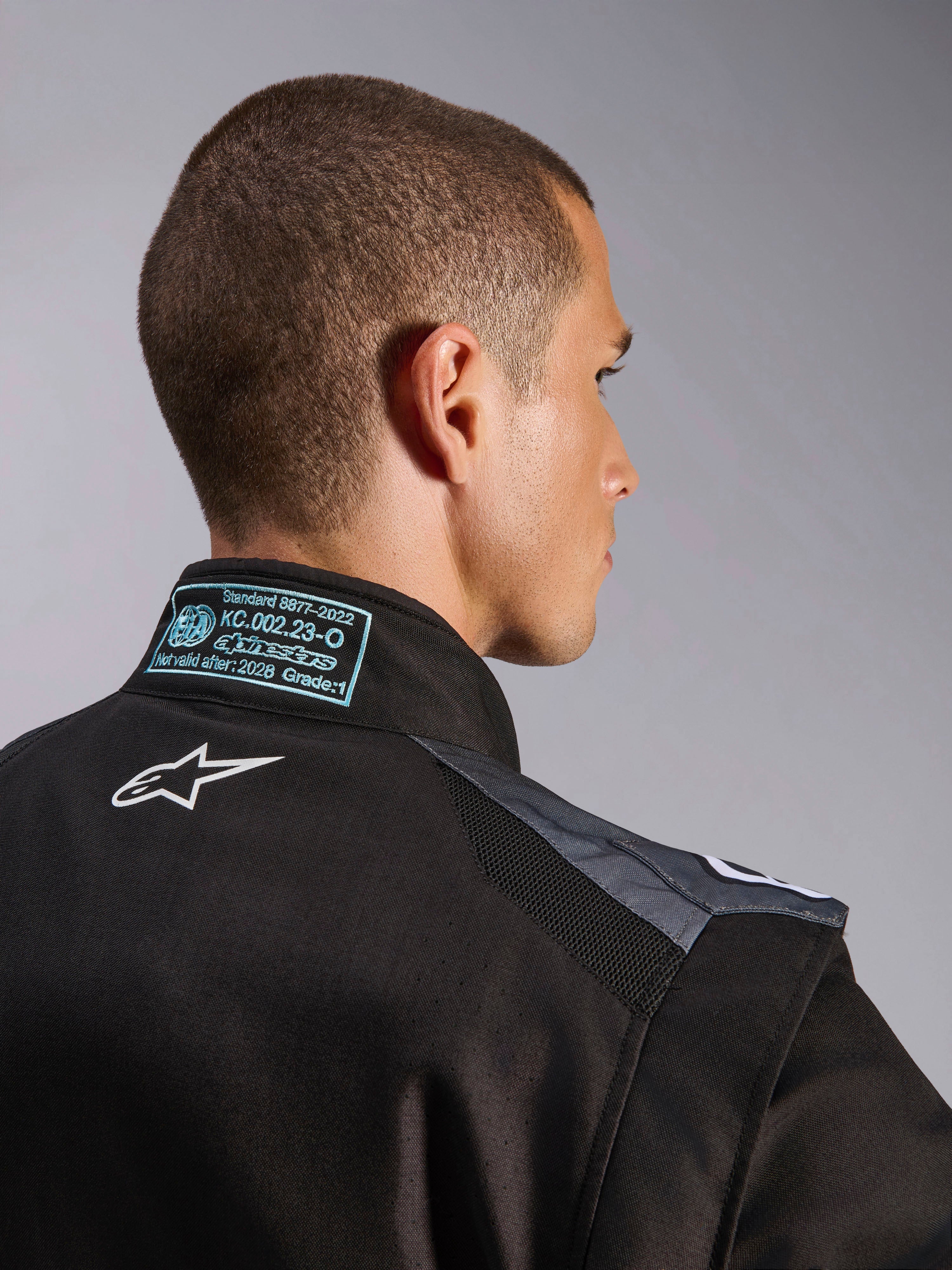 Macacão Alpinestars KMX-5 V3 em Preto e Dark Shadow, vista detalhada das costas e da gola com o logótipo Astars branco, etiqueta de certificação FIA 8877-2022 Grau 1 e painéis de malha ventilada nos ombros.
