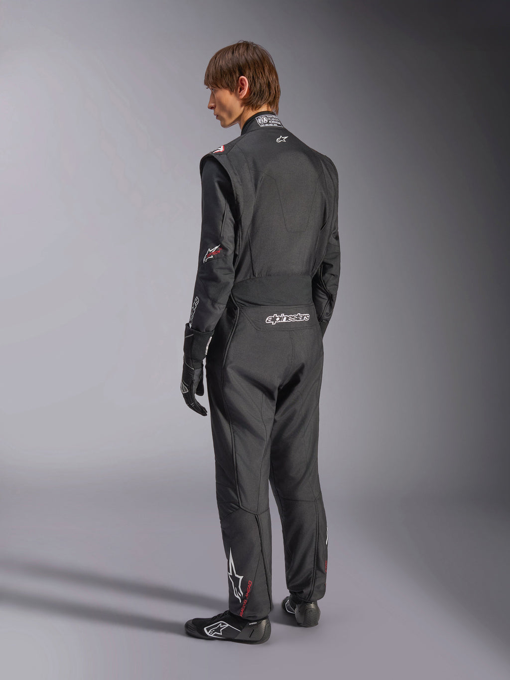 Macacão Alpinestars GP Tech V4, fato de automobilismo, preto com detalhes brancos, vista traseira de modelo a usar o fato com luvas e sapatos de competição, com logótipos brancos na zona lombar e na perna, equipamento de segurança profissional para desportos motorizados