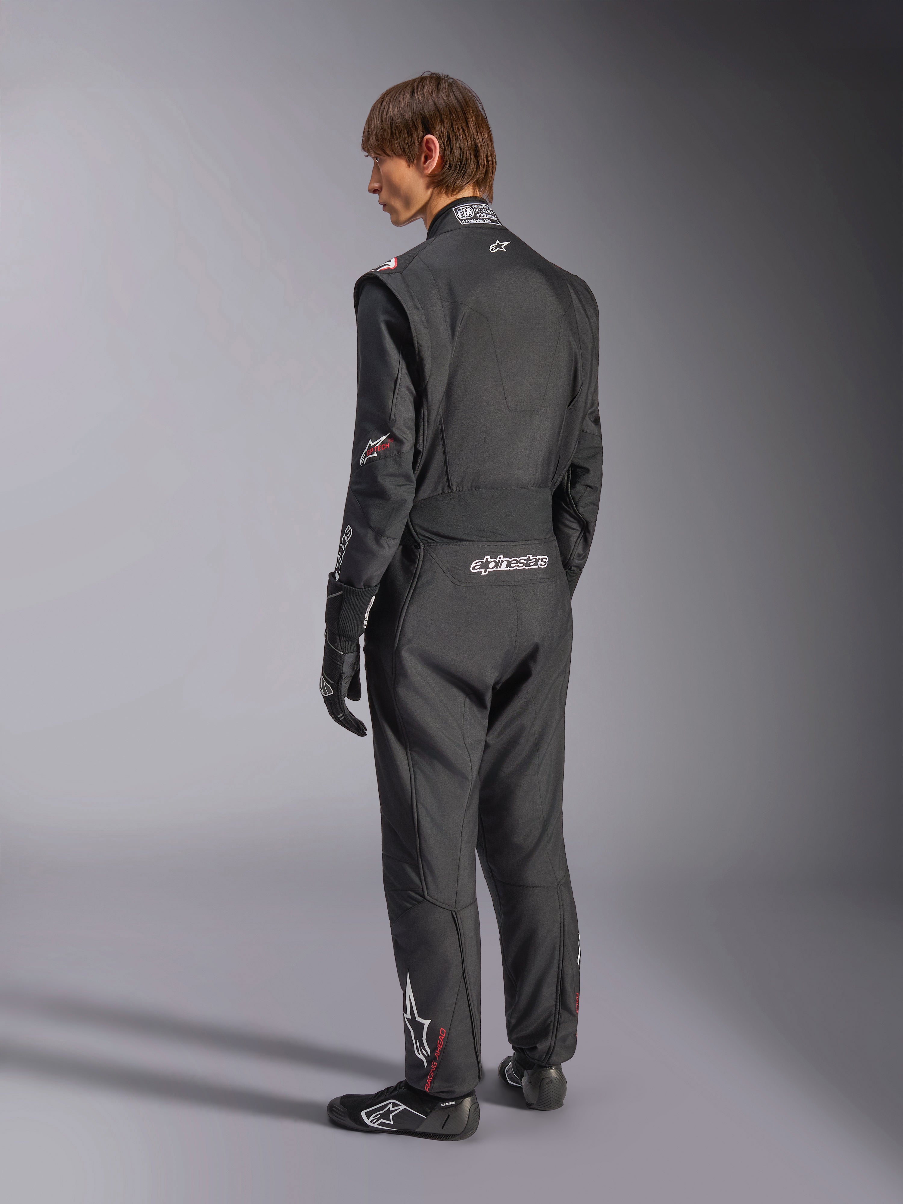 Macacão Alpinestars GP Tech V4, fato de automobilismo, preto com detalhes brancos, vista traseira de modelo a usar o fato com luvas e sapatos de competição, com logótipos brancos na zona lombar e na perna, equipamento de segurança profissional para desportos motorizados