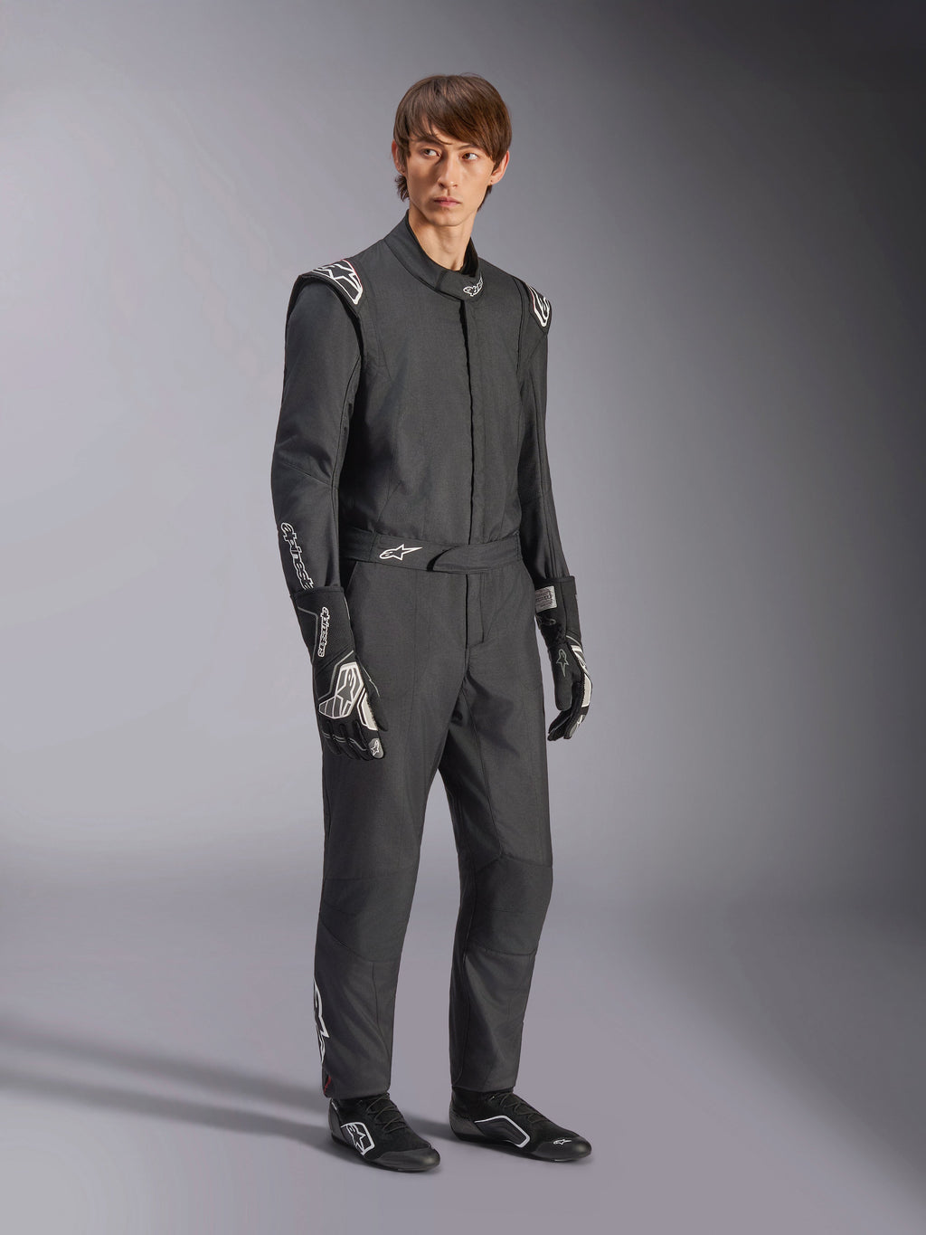 Macacão Alpinestars GP Tech V4, macacão de automobilismo de uma peça, preto e branco, usado com luvas e calçado de competição a combinar, com logótipos brancos nos ombros e antebraços, design profissional e elegante para desportos motorizados