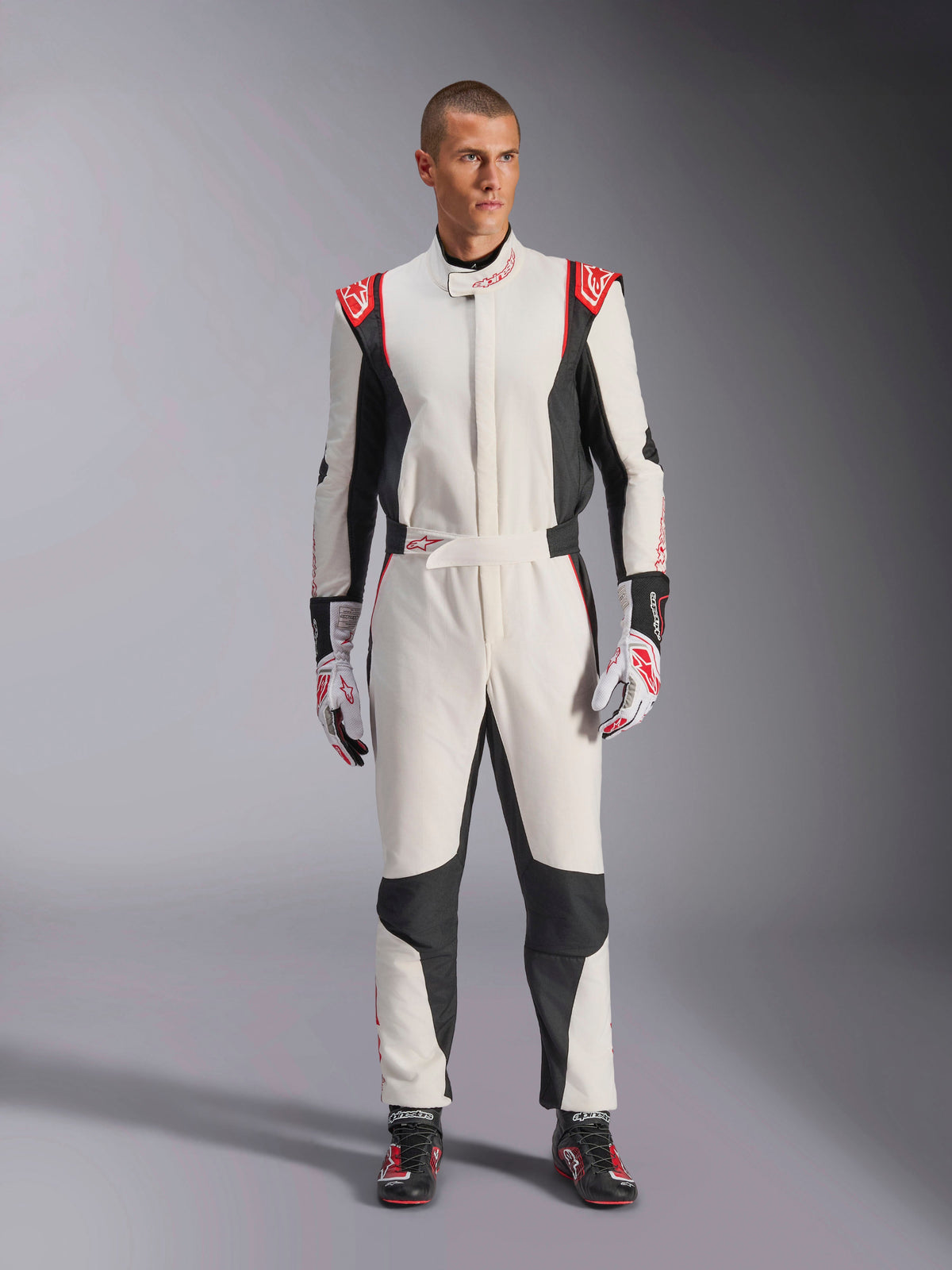 Macacão Alpinestars GP Tech V4, macacão de automobilismo branco, preto e vermelho, vestido por um modelo masculino com luvas e sapatos de corrida a condizer, com logótipos vermelhos nos ombros e painéis laterais pretos, equipamento profissional de desportos motorizados para uso em pista