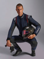 Piloto de corridas masculino vestindo o Macacão Alpinestars GP Tech V4 em azul-marinho com detalhes pretos e vermelhos, segurando um capacete de fibra de carbono com viseira iridescente, luvas e botas de corrida pretas, equipamento profissional de automobilismo