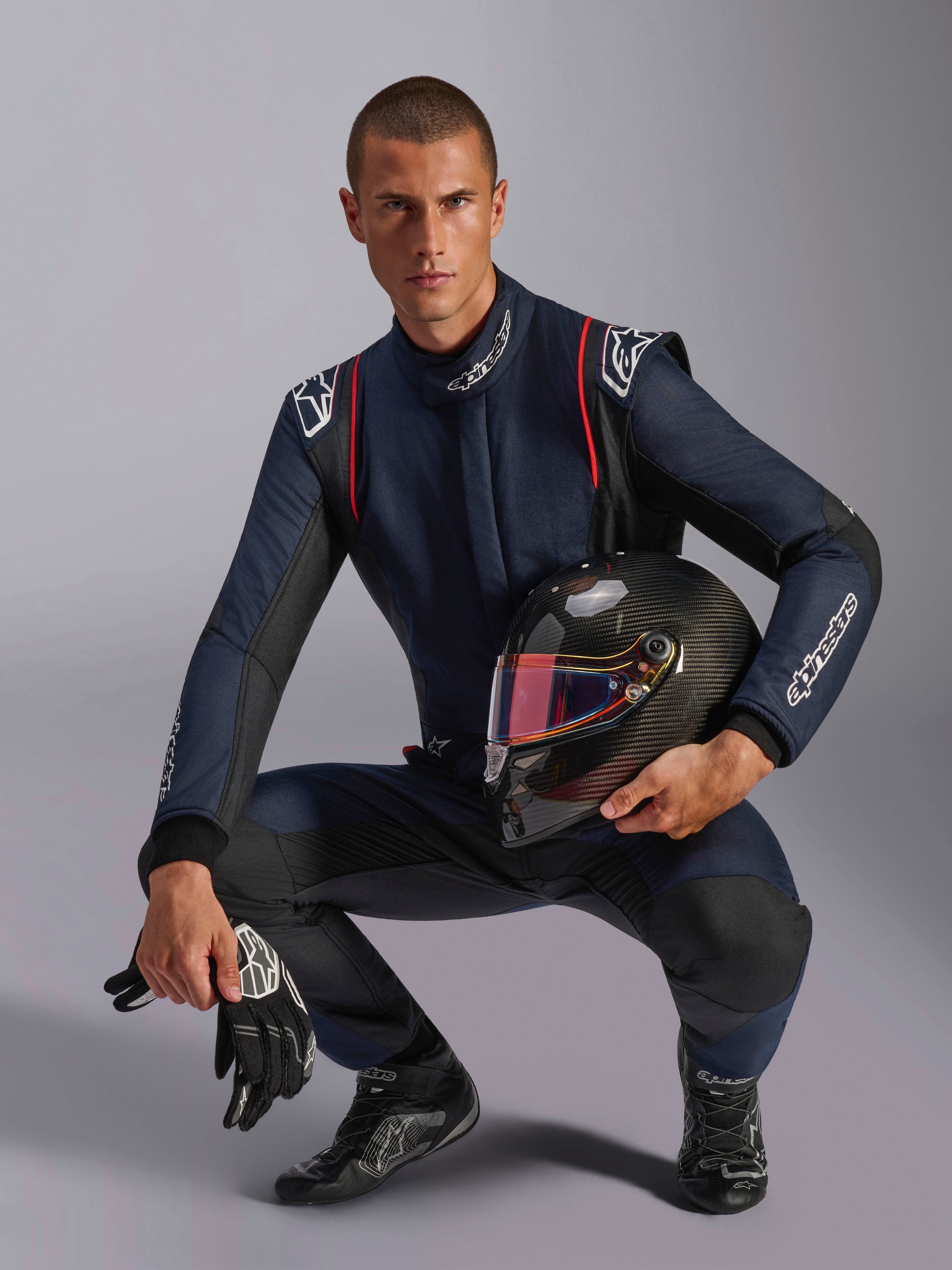 Piloto de corridas masculino vestindo o Macacão Alpinestars GP Tech V4 em azul-marinho com detalhes pretos e vermelhos, segurando um capacete de fibra de carbono com viseira iridescente, luvas e botas de corrida pretas, equipamento profissional de automobilismo