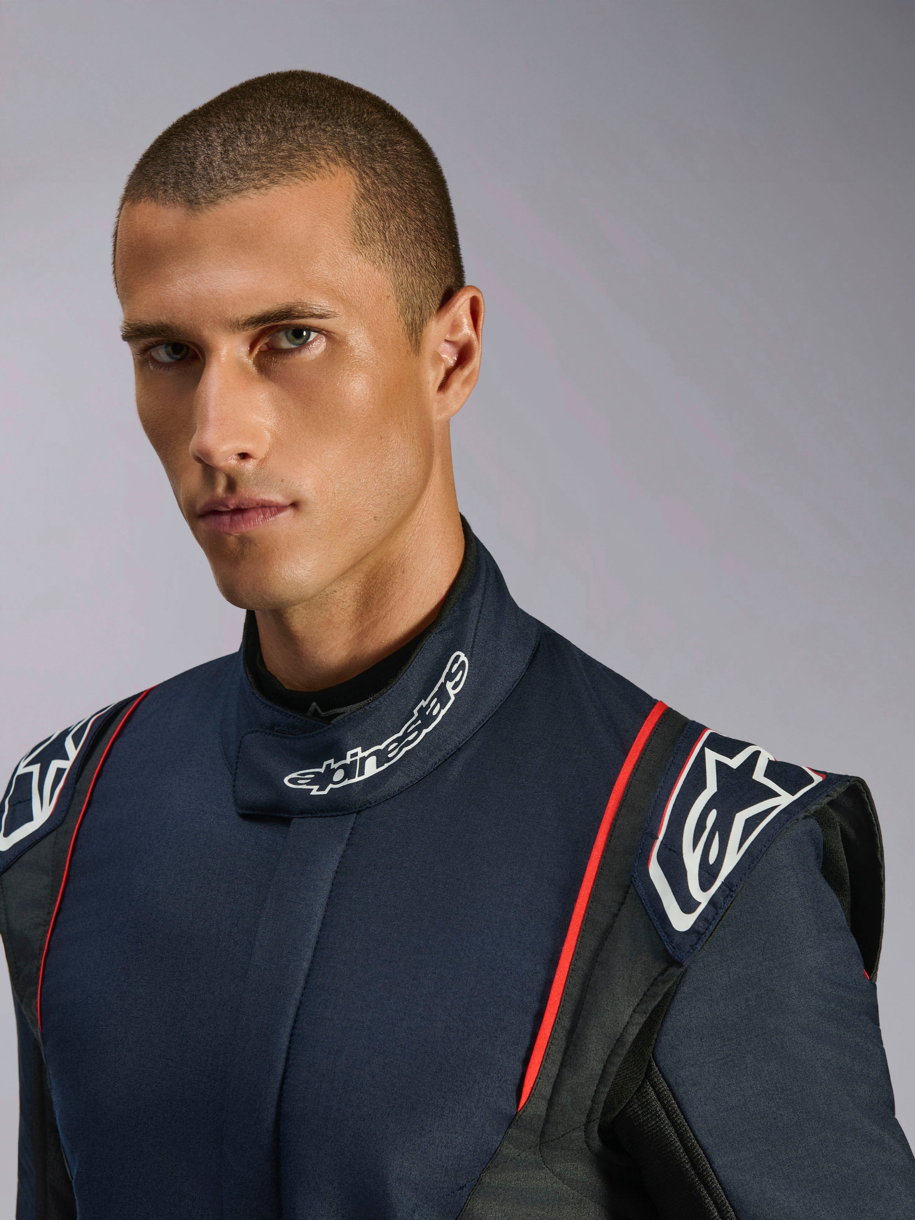 Retrato em grande plano de um modelo a usar o Macacão Alpinestars GP Tech V4, macacão de automobilismo em azul-marinho, preto e vermelho, com a marca Alpinestars na gola alta e ombreiras estruturadas.
