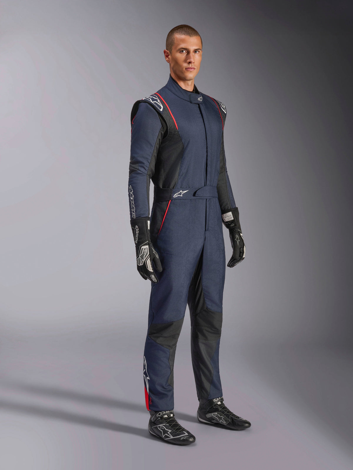 Macacão Alpinestars GP Tech V4, macacão de automobilismo de uma peça, azul marinho, preto e vermelho, vestido por um modelo com luvas e botas de corrida pretas, com painéis pretos e vivos vermelhos, equipamento profissional de desporto motorizado