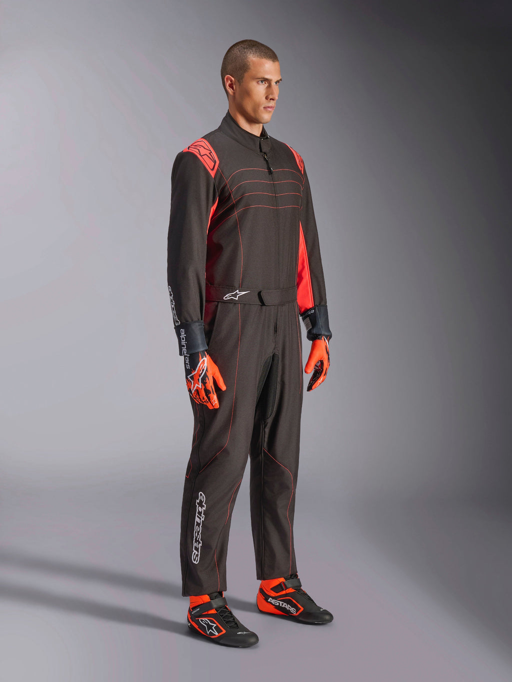 Fato Alpinestars KMX-9 V3, fato de karting, preto e vermelho fluorescente, vestido por um modelo com luvas cor-de-laranja e sapatos de corrida pretos, com costuras vermelhas, detalhes nos ombros e logótipos brancos, design atlético para competição profissional