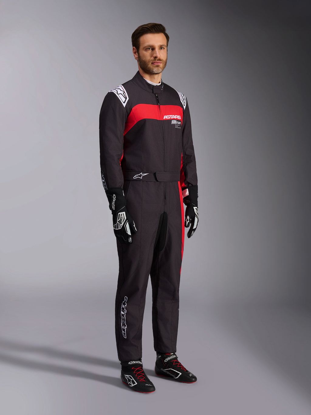 Modelo masculino a usar o fato Alpinestars KMX-9 V3 Graphic 3, fato de karting, preto e vermelho, com dragonas brancas nos ombros e logótipo vermelho no peito, combinado com luvas de corrida pretas e sapatos com atacadores vermelhos, vestuário profissional de desporto motorizado