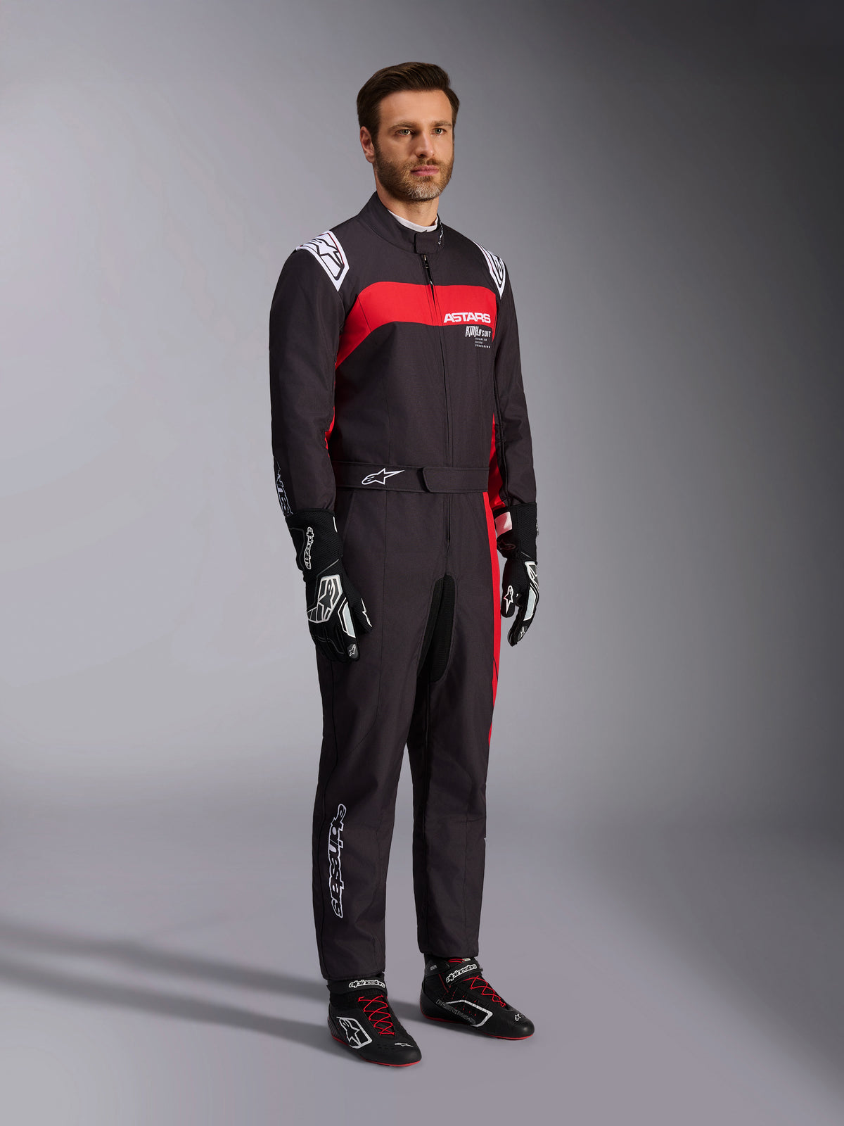 Modelo masculino a usar o fato Alpinestars KMX-9 V3 Graphic 3, fato de karting, preto e vermelho, com dragonas brancas nos ombros e logótipo vermelho no peito, combinado com luvas de corrida pretas e sapatos com atacadores vermelhos, vestuário profissional de desporto motorizado