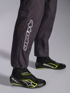 Alpinestars KMX-9 V3 Graphic 3 Suit, macacão de karting, preto, grande plano da parte inferior das pernas e dos punhos dos tornozelos, com a marca Alpinestars vertical em branco, combinado com sapatos de karting pretos e amarelo fluorescente, pormenor de vestuário de karting profissional.