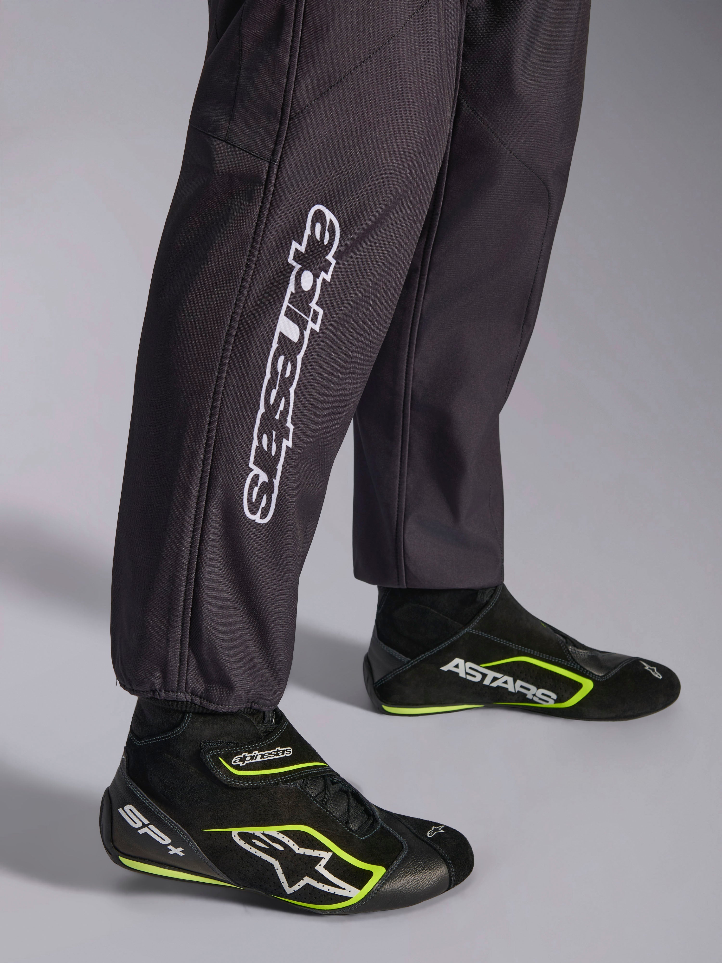 Alpinestars KMX-9 V3 Graphic 3 Suit, macacão de karting, preto, grande plano da parte inferior das pernas e dos punhos dos tornozelos, com a marca Alpinestars vertical em branco, combinado com sapatos de karting pretos e amarelo fluorescente, pormenor de vestuário de karting profissional.