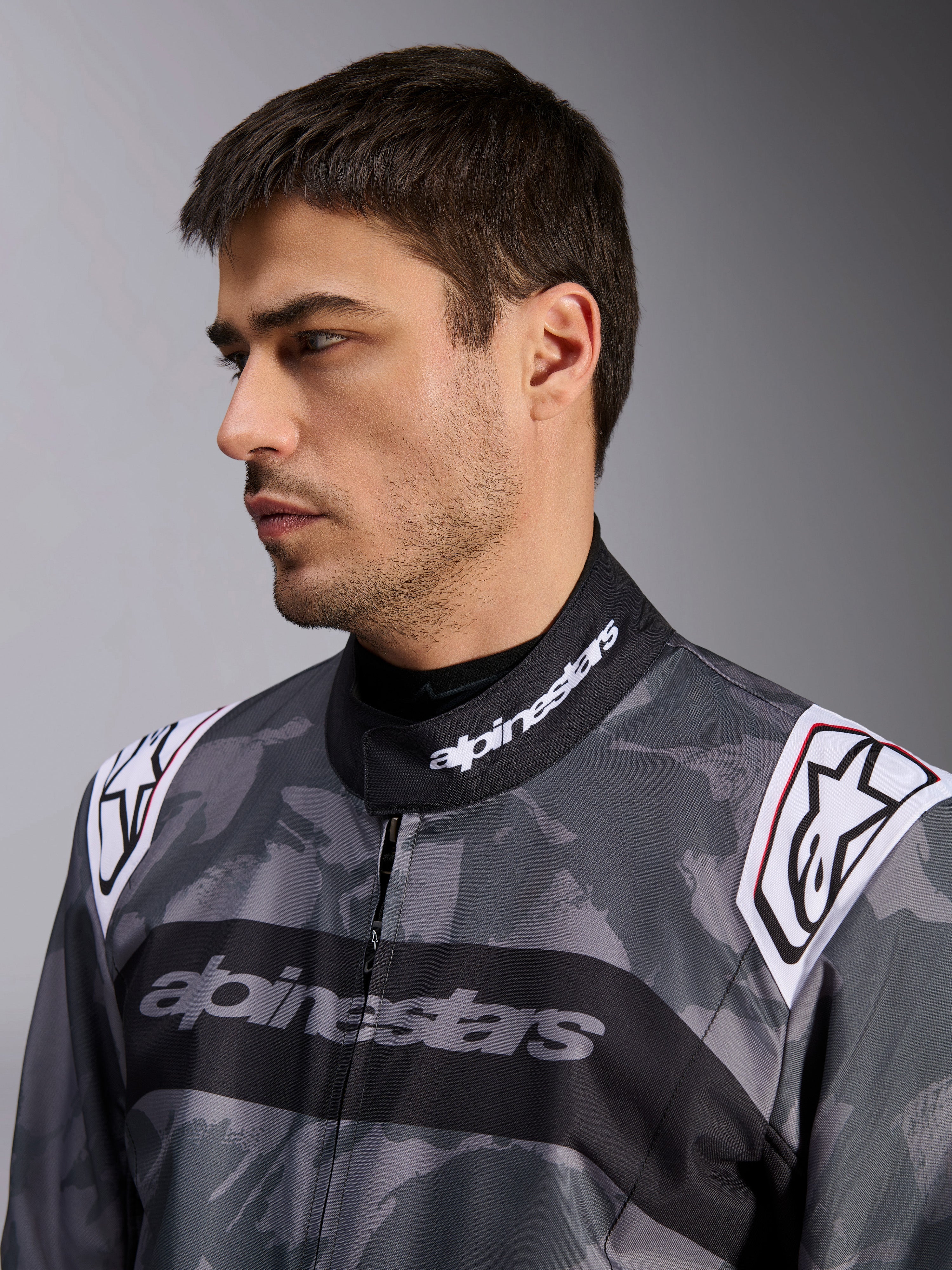 Alpinestars KMX-9 V3 Graphic 5 Suit, macacão de karting, padrão de camuflagem cinzento escuro e preto, grande plano da gola e ombros com logótipos brancos, vestuário de corrida profissional