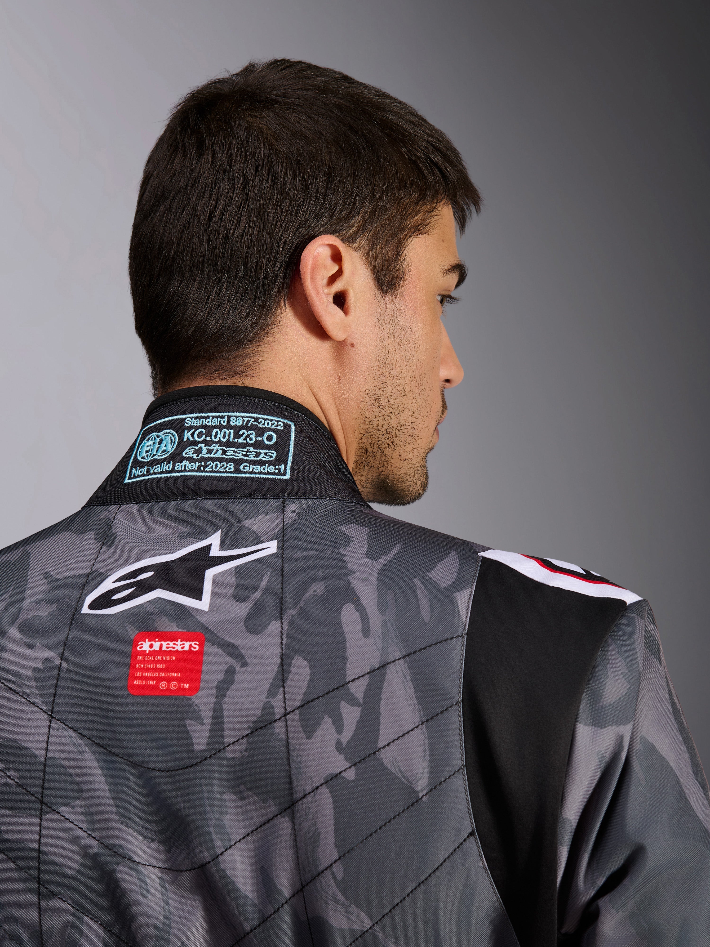 Macacão Alpinestars KMX-9 V3 Graphic 5, fato de karting, padrão de camuflagem em cinzento escuro e preto, pormenor da parte superior das costas mostrando o logótipo Alpinestars, etiqueta de marca vermelha e selo de homologação FIA no colarinho.