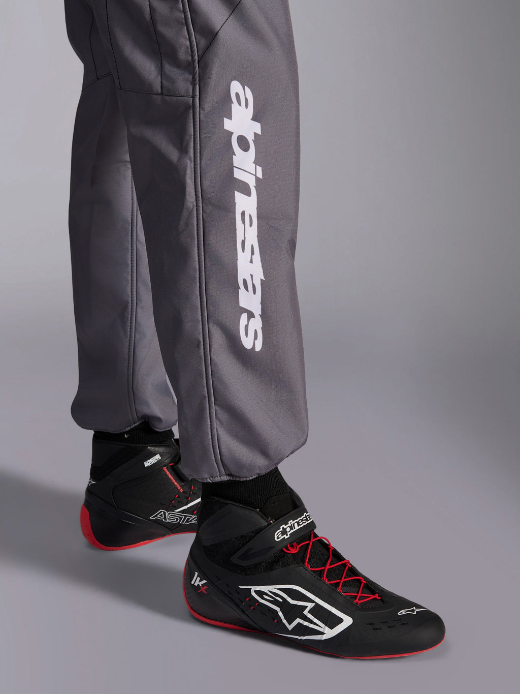Macacão Alpinestars KMX-9 V3 Graphic 5, fato de karting, cinzento escuro, pormenor da parte inferior da perna com logótipo vertical Alpinestars em branco, usado com botas de corrida pretas e vermelhas, material durável para competição de karting