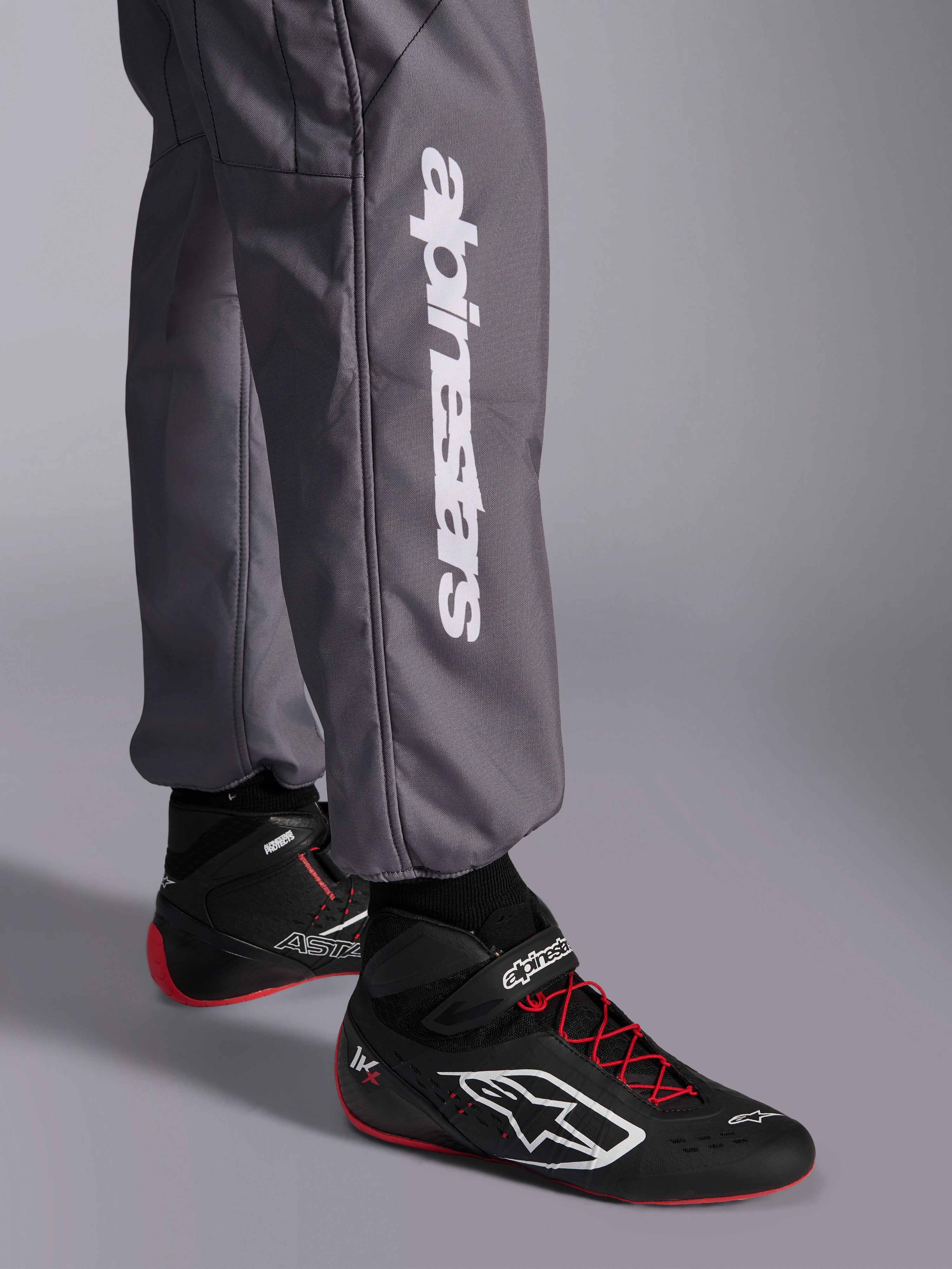 Macacão Alpinestars KMX-9 V3 Graphic 5, fato de karting, cinzento escuro, pormenor da parte inferior da perna com logótipo vertical Alpinestars em branco, usado com botas de corrida pretas e vermelhas, material durável para competição de karting