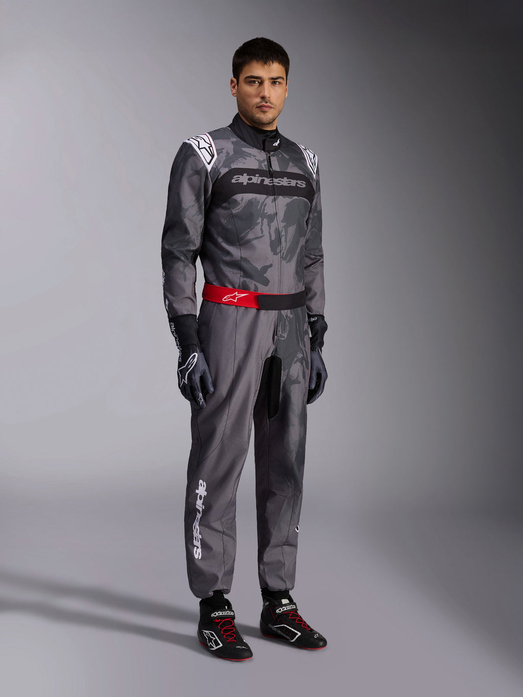 Fato Alpinestars KMX-9 V3 Graphic 5, fato de karting, cinzento escuro e preto com padrão gráfico camuflado, cinto vermelho com logótipo branco, usado com luvas e botas de corrida, equipamento profissional de desporto motorizado para karting