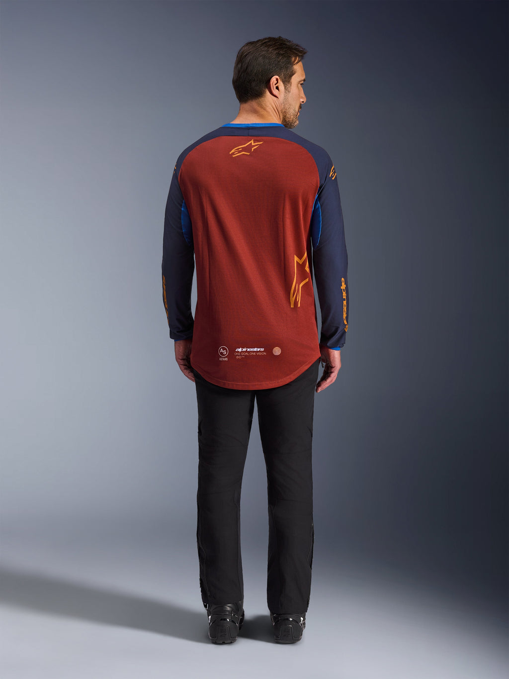 Alpinestars Maxdura Dual Jersey em Rust Brown e Mood Indigo, vista traseira mostrando a bainha traseira descaída e os detalhes do logótipo, usada com calças técnicas e botas pretas, concebida para motociclismo de aventura e dual-sport.