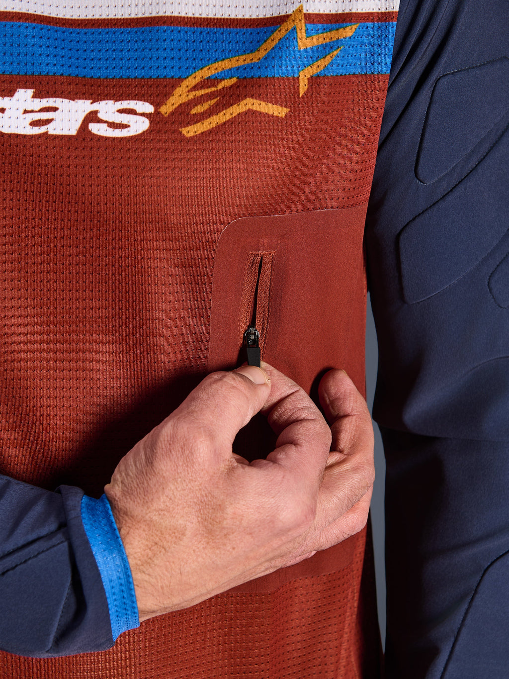 Primeiro plano da Alpinestars Maxdura Dual Jersey, torso em malha Rust Brown com bolso no peito com fecho de correr sem costuras, mangas Mood Indigo com acolchoamento integrado, pormenor de camisola técnica de motociclismo de aventura