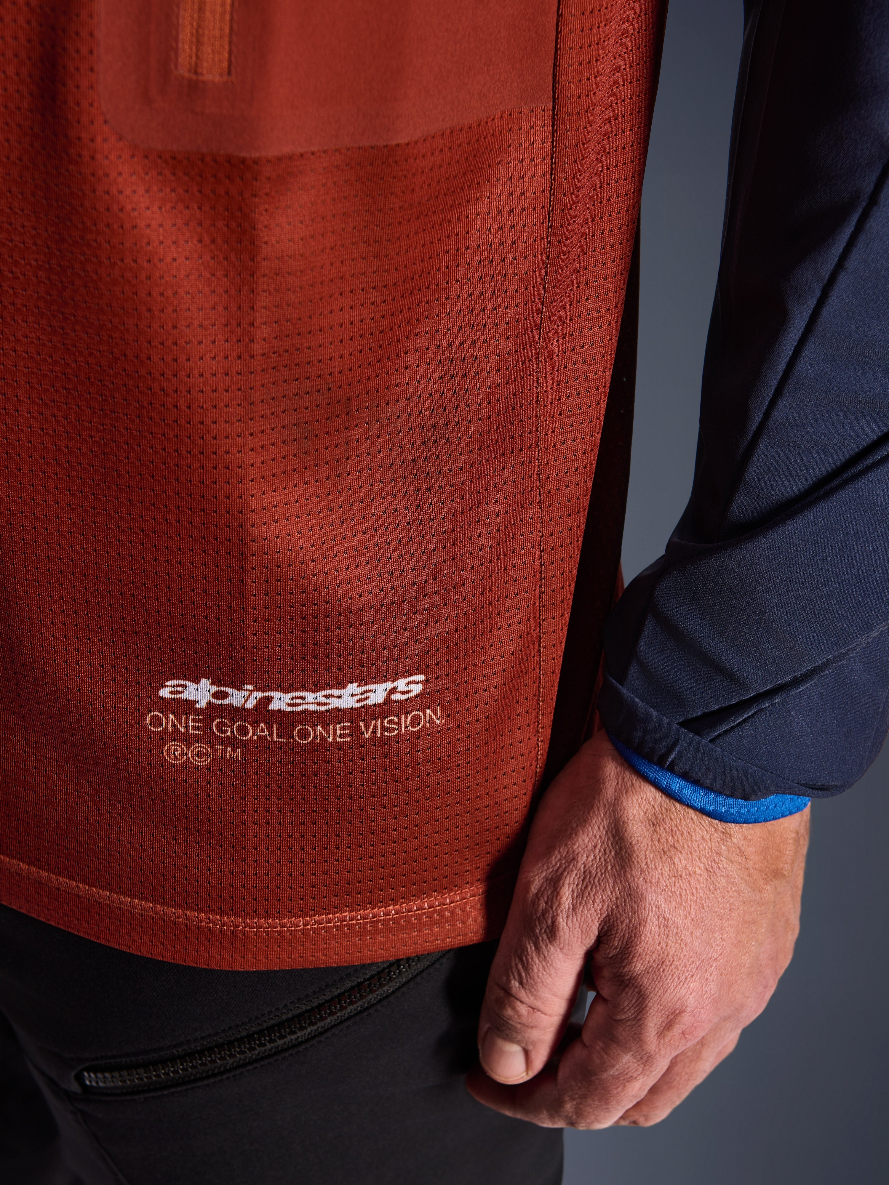 Alpinestars Maxdura Dual Jersey, camisola de motociclismo de aventura, castanho ferrugem e azul Mood Indigo, vista em pormenor do tecido em malha do tronco e da manga tecida em azul-marinho, logótipo Alpinestars 'One Goal One Vision' a branco na bainha inferior.