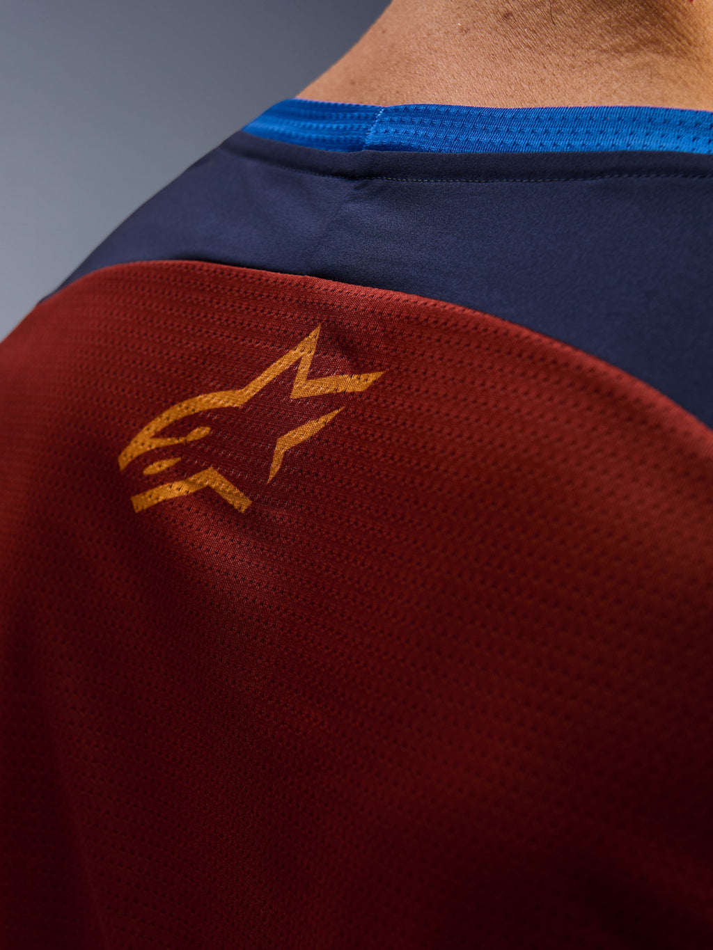Primeiro plano da Alpinestars Maxdura Dual Jersey, tecido de malha castanho ferrugem nas costas, pequeno logótipo de estrela cor de laranja, painel superior azul escuro e rebordo da gola em azul vivo, vista detalhada de camisola de motociclismo de aventura