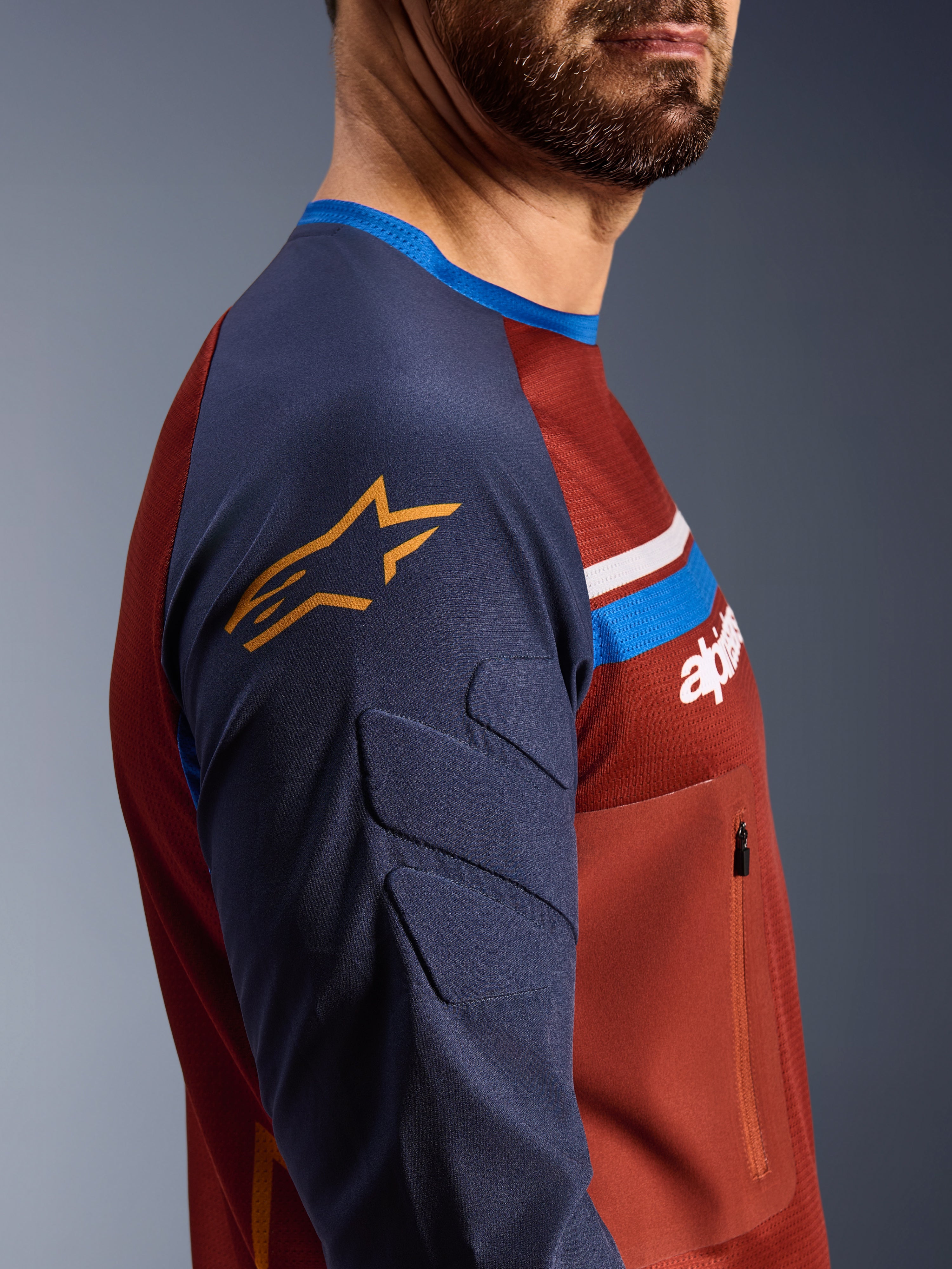 Alpinestars Maxdura Dual Jersey, Castanho Ferrugem e Azul Mood Indigo, vista de perfil lateral mostrando mangas acolchoadas com logótipo de estrela amarelo, construção do tronco em malha com bolso no peito com fecho de correr, vestuário técnico de motociclismo para condução de aventura