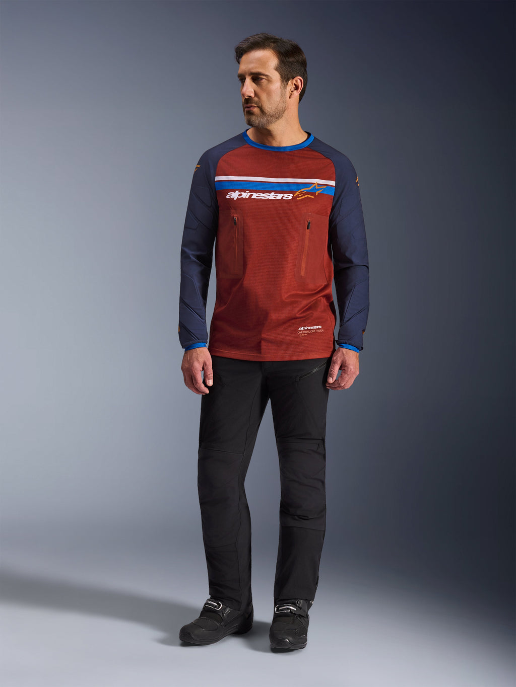 Alpinestars Maxdura Dual Jersey, Rust Brown e Mood Indigo, camisola de motociclismo de aventura com tronco em malha e mangas azul-marinho, usada por um modelo com calças de condução pretas e botas pretas, vestuário técnico dual-sport para condução off-road