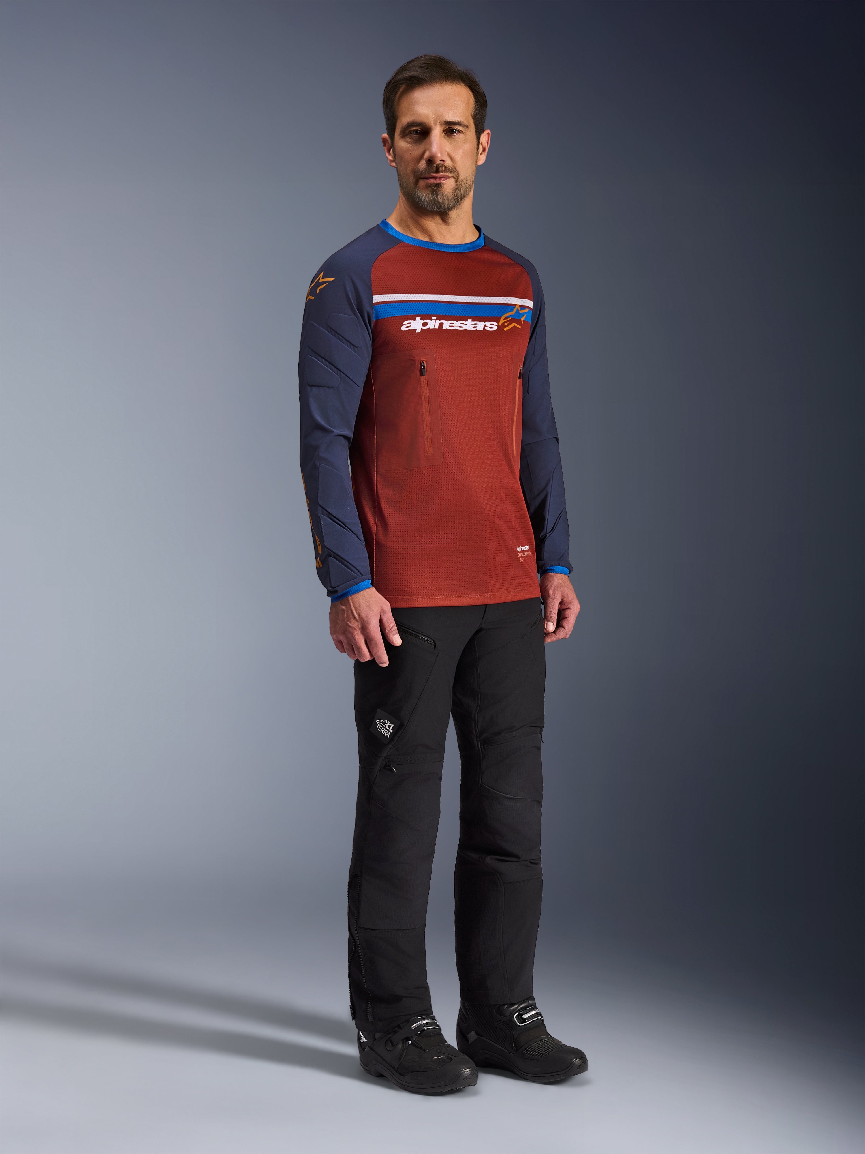 Homem a usar a Alpinestars Maxdura Dual Jersey em Rust Brown e Mood Indigo, com tronco em malha e mangas acolchoadas, calças e botas de motociclismo pretas, vestuário de aventura para motociclismo