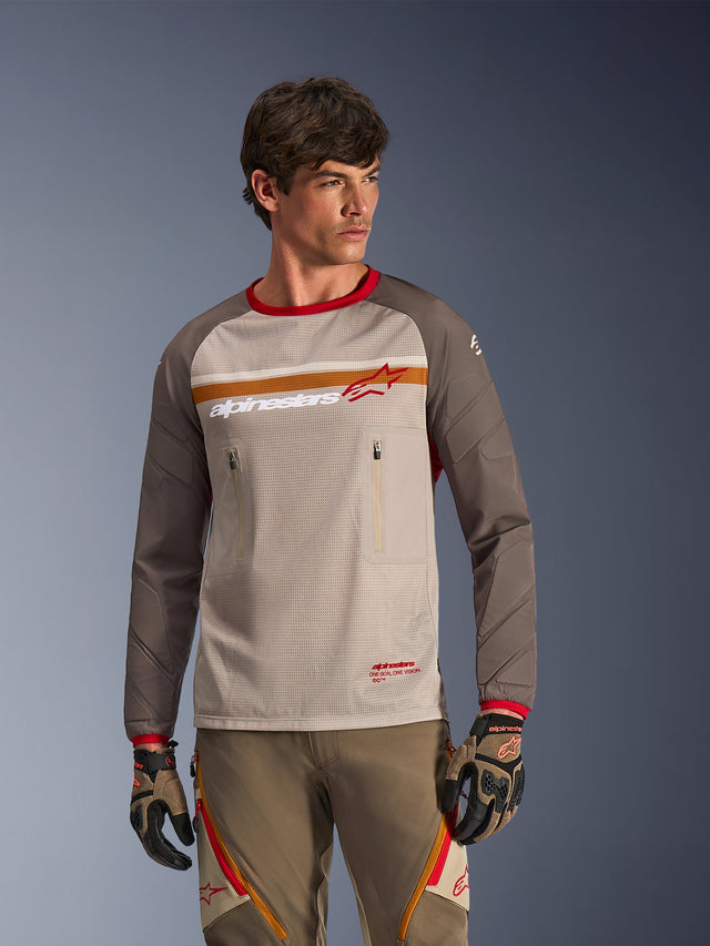 Alpinestars Maxdura Dual Jersey em Aluminum Walnut, camisola de motociclismo de aventura com torso em malha respirável, mangas castanho nogueira resistentes à abrasão e bolsos no peito com fecho de correr, vestida por um modelo com equipamento off-road a condizer.